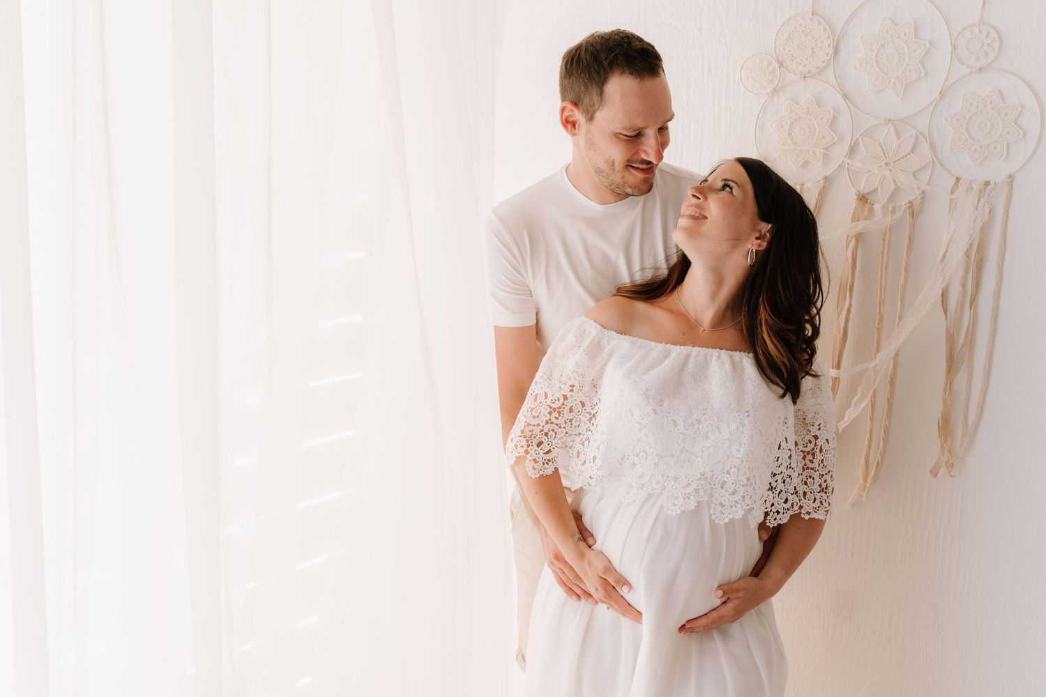 Couple embracing in bright boho studio, man holding pregnant woman's belly lovingly.