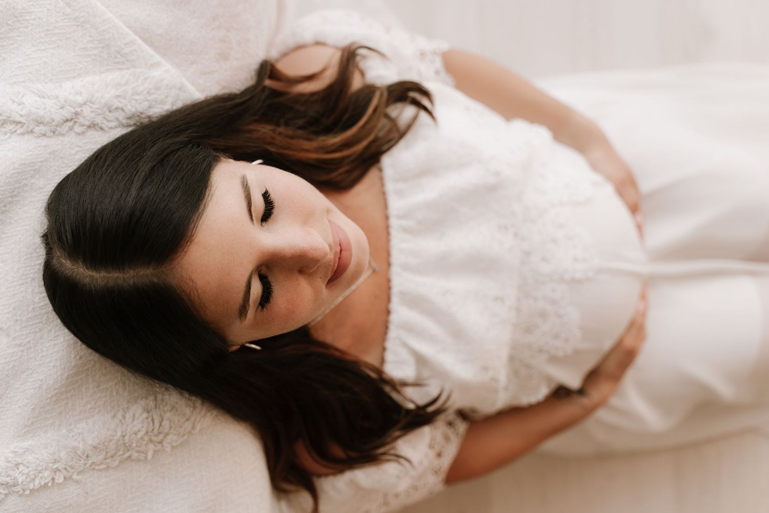 Young woman with dark hair lying on white bedding wearing a delicate lace dress, eyes closed.