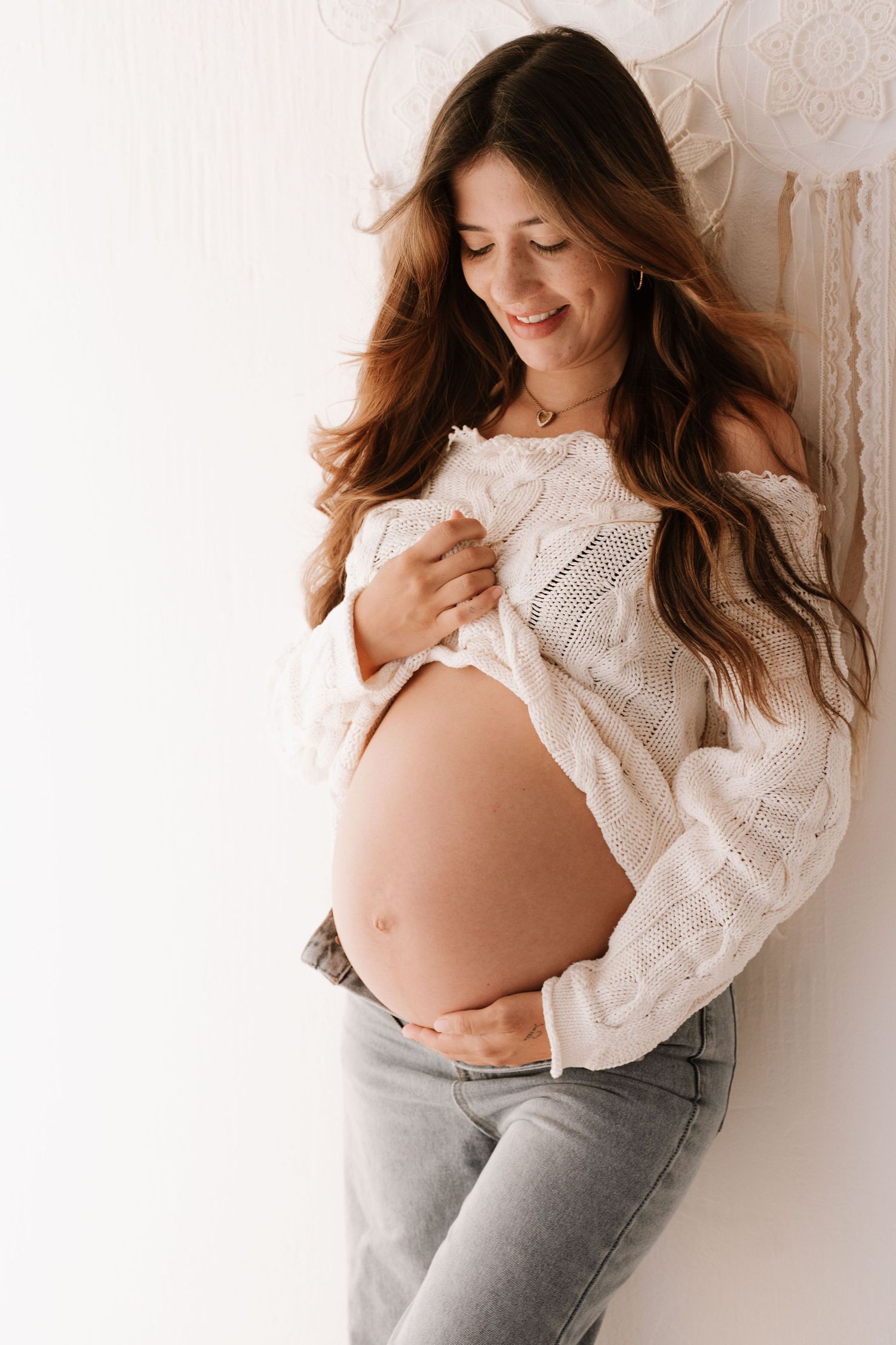 Smiling pregnant woman in open knit sweater and jeans showing bare baby bump.