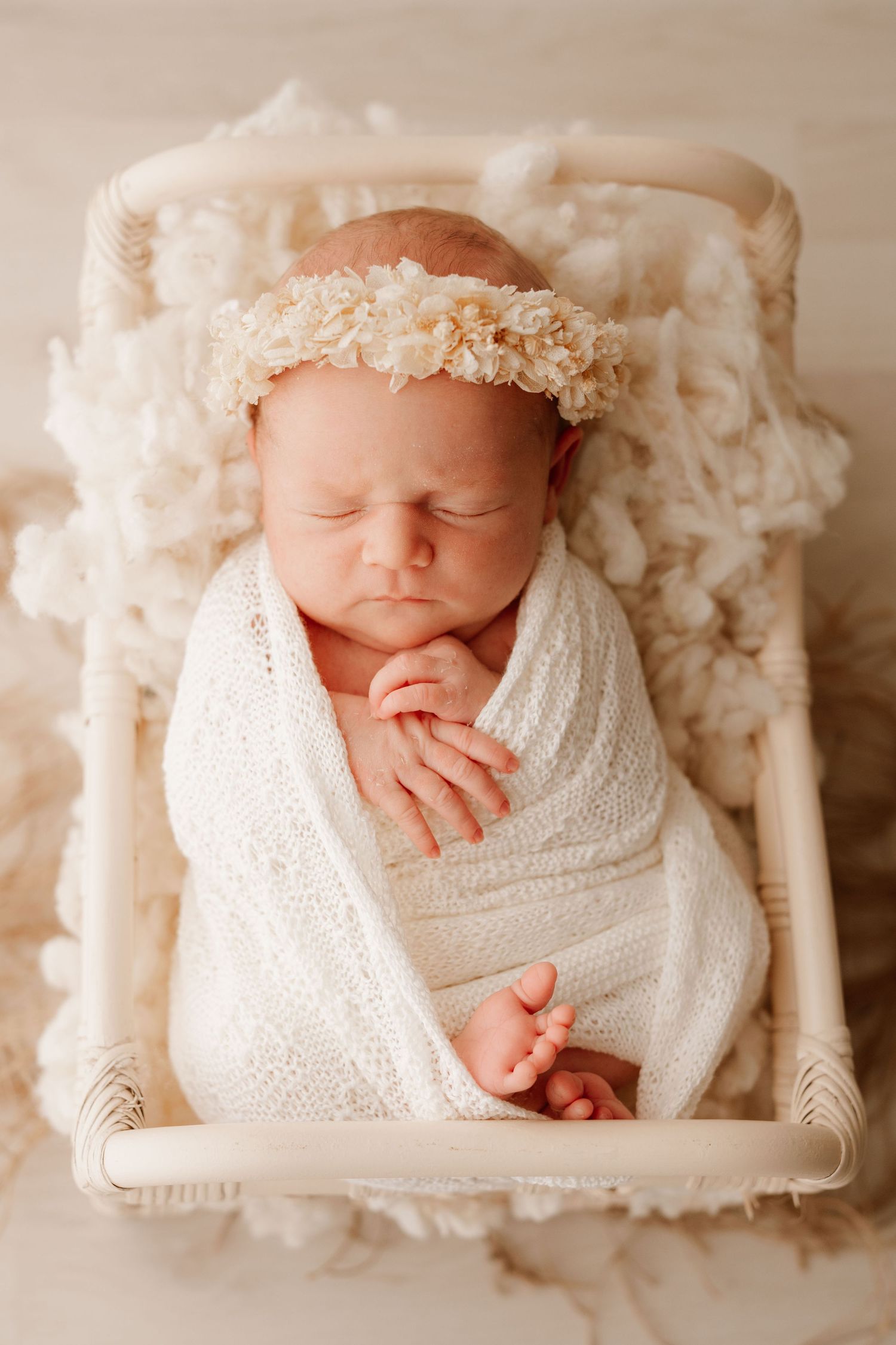 Sleeping newborn baby swaddled in white wrap with floral headband, resting in a small cream wooden bed.
