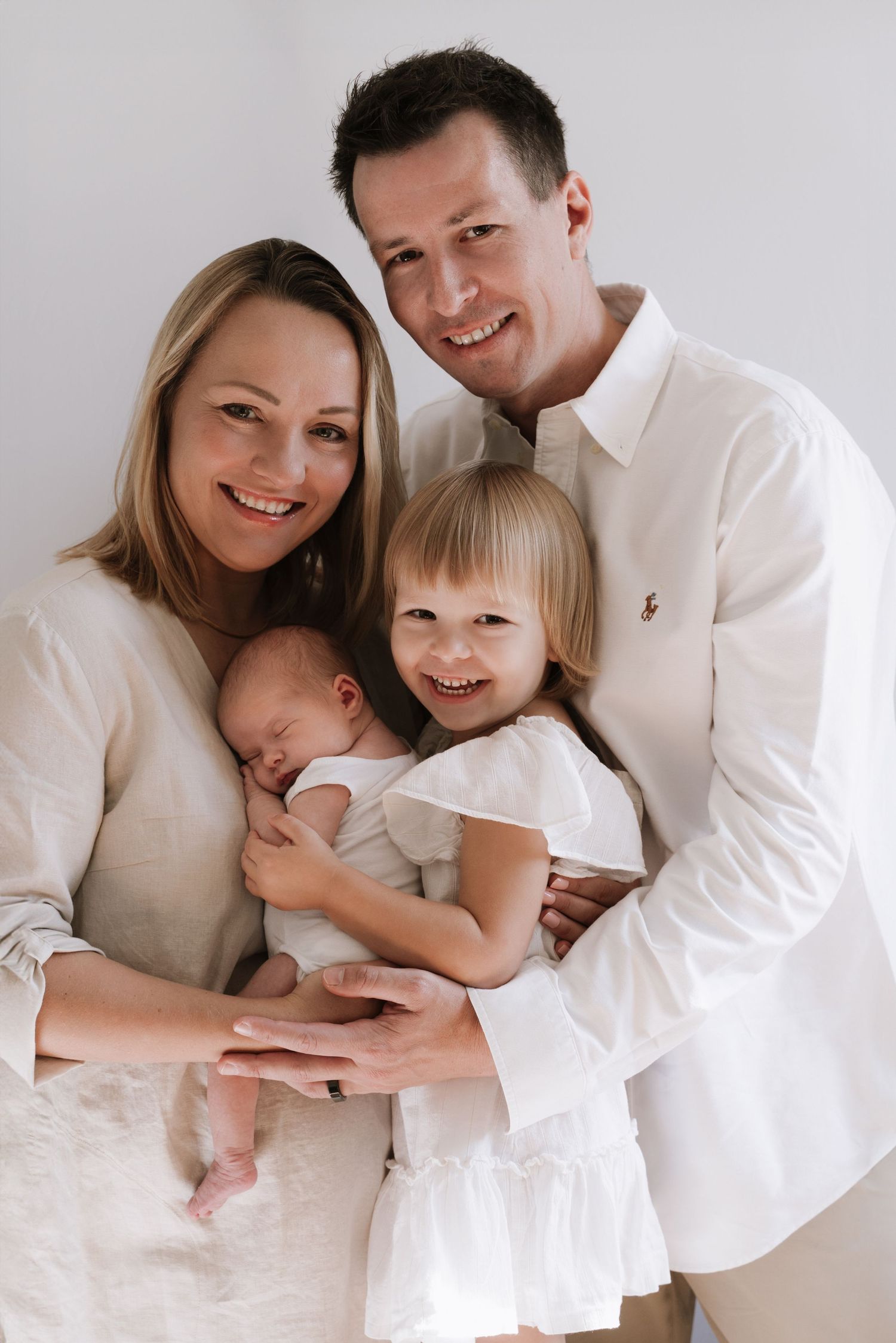 Happy family of four in white outfits posing with newborn baby and toddler daughter in studio.