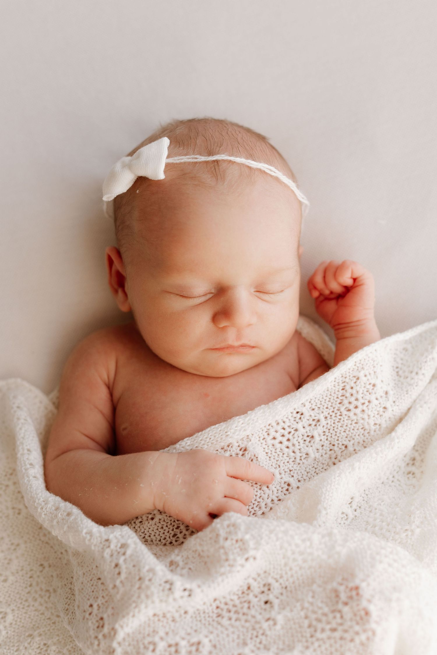 Sleeping newborn baby girl wrapped in white lace blanket wearing a delicate white bow headband.