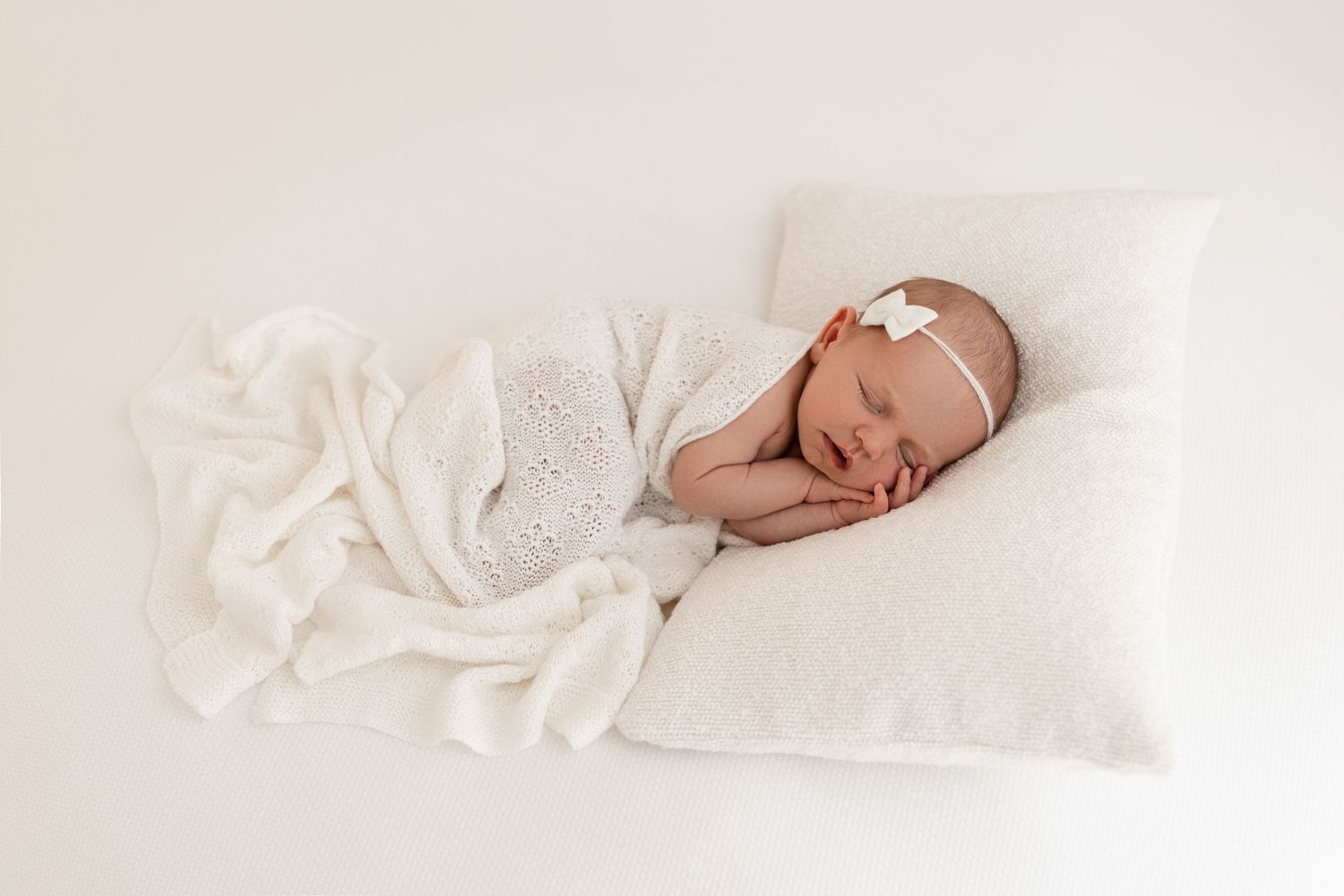 Sleeping newborn baby wrapped in white blanket on fluffy pillow with delicate bow headband, white background.