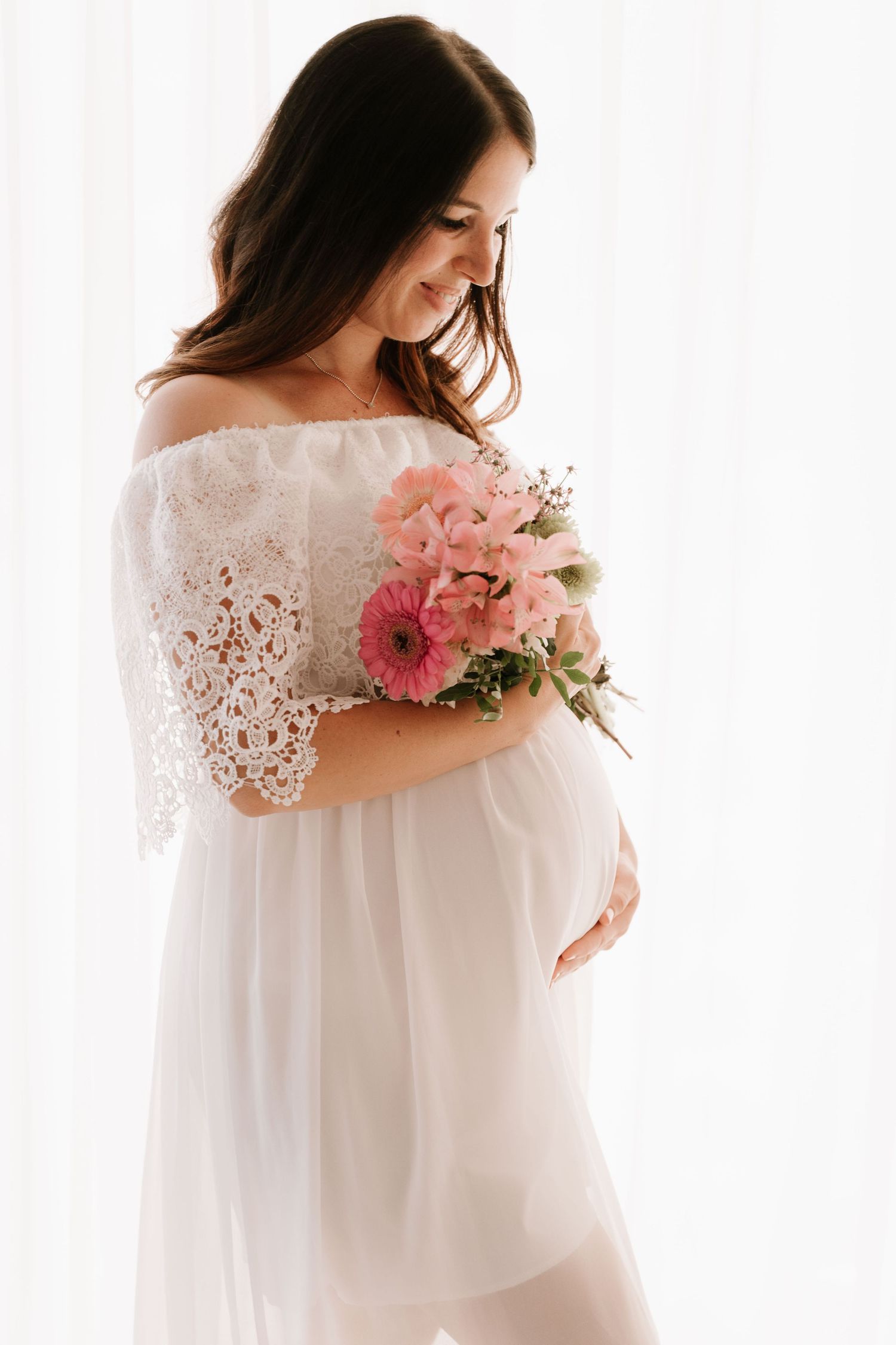 Smiling pregnant woman in white lace dress cradling belly while holding pink flowers.