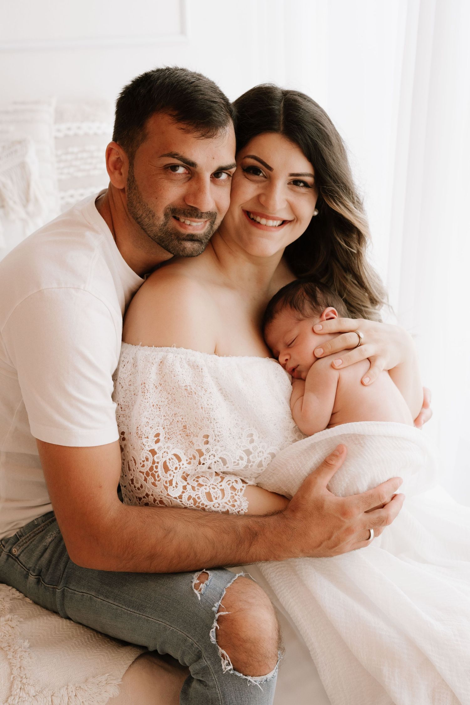 A happy couple in white outfits tenderly holds their sleeping newborn baby in a bright, airy studio setting.