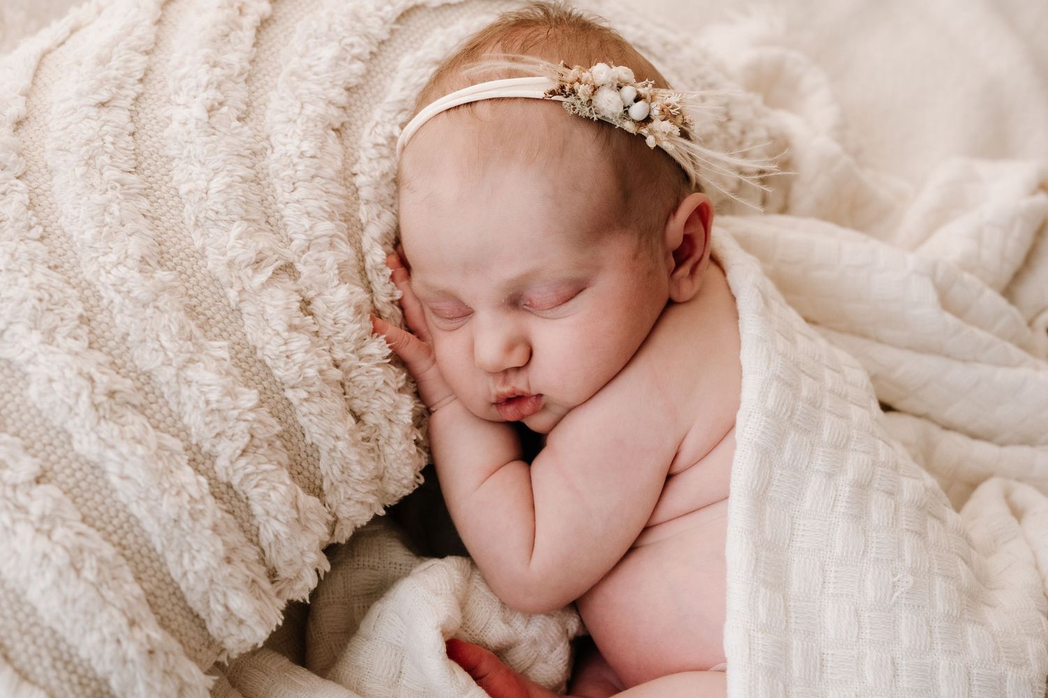 Peaceful sleeping newborn baby girl with floral headband wrapped in cream textured blanket.