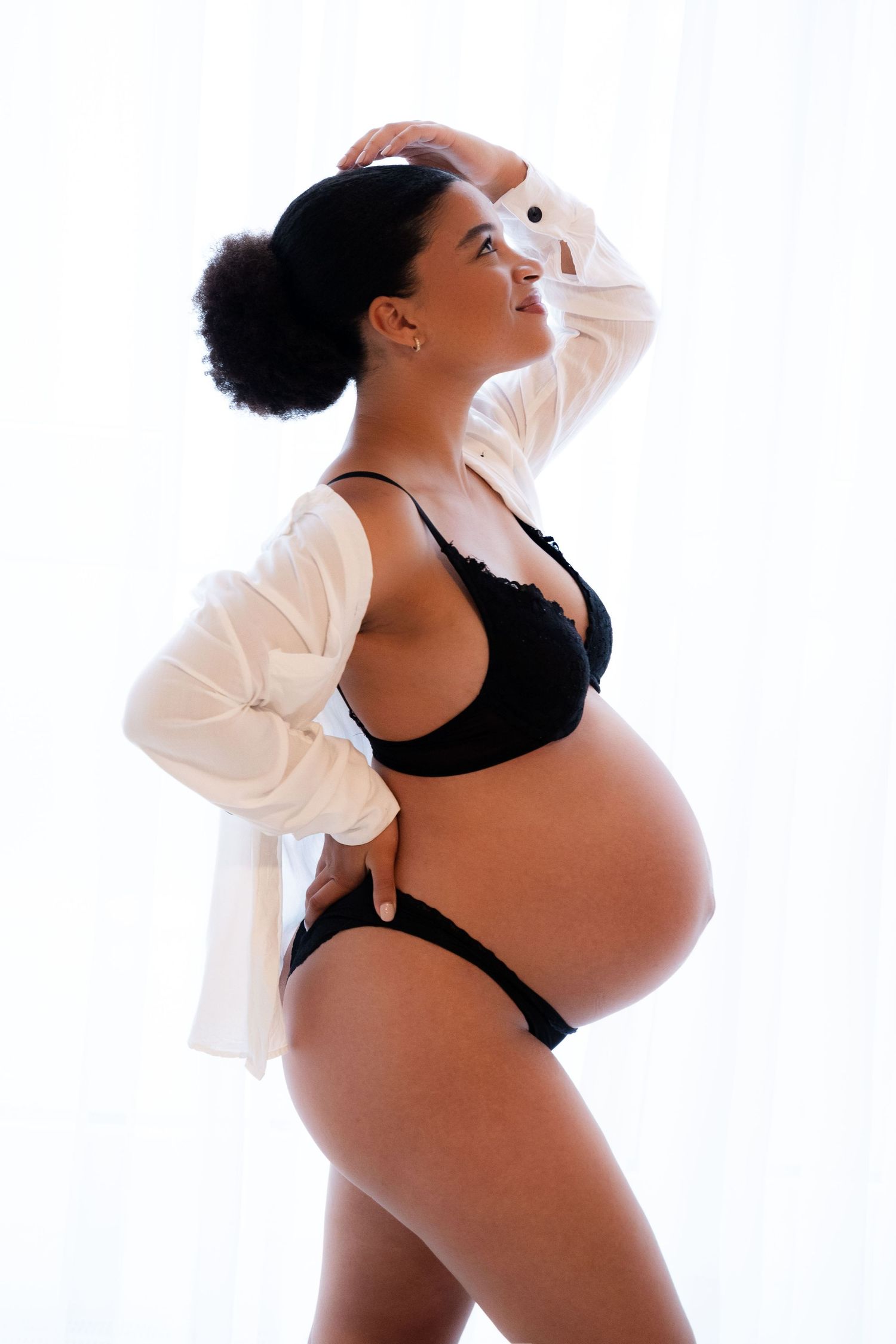 Pregnant woman in black lingerie and open white shirt posing sideways, showing baby bump.