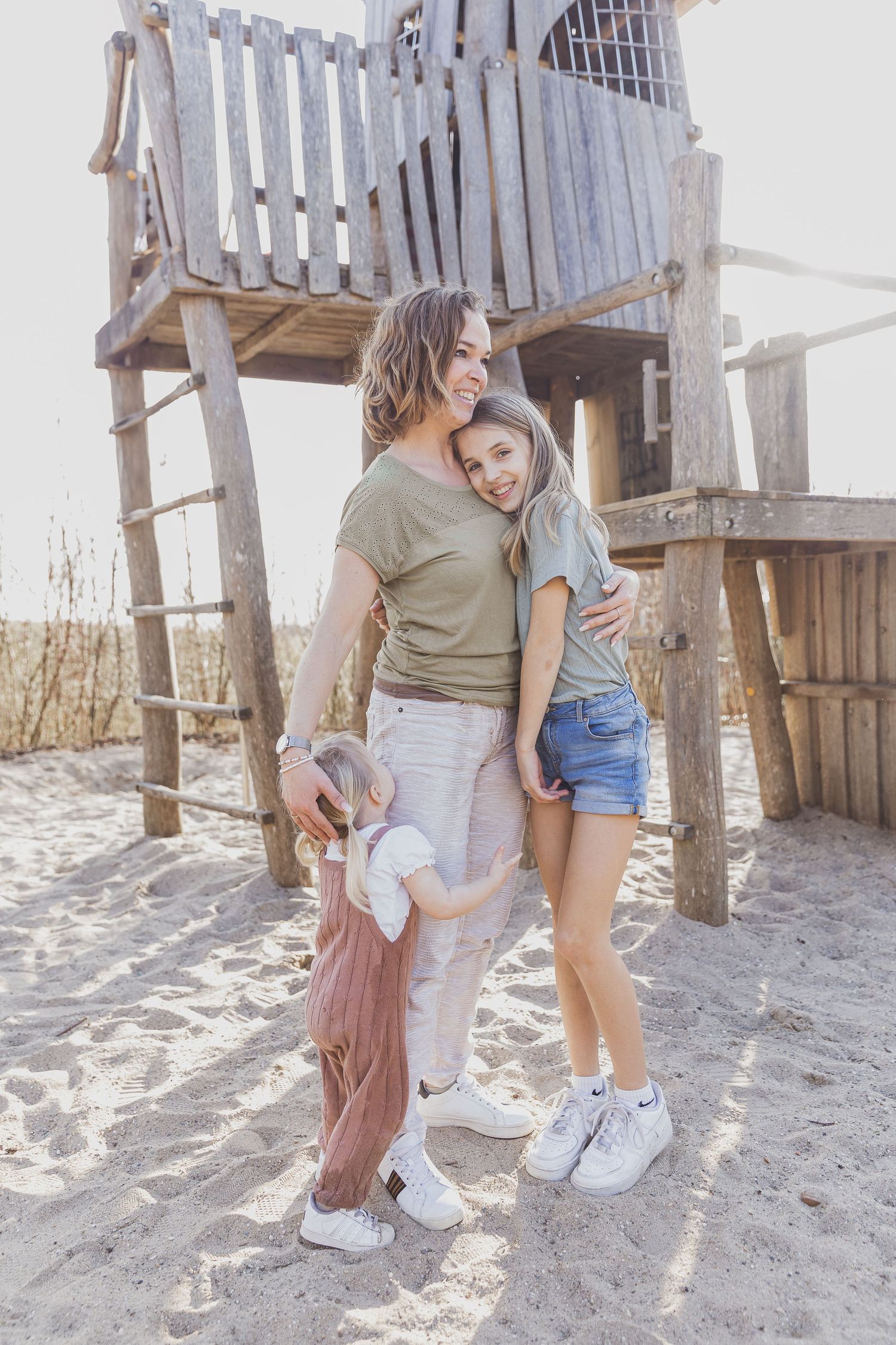 People sharing a tender moment by a wooden playground structure on a beach. Gezinsfotografie Nederland, ongewdongen.