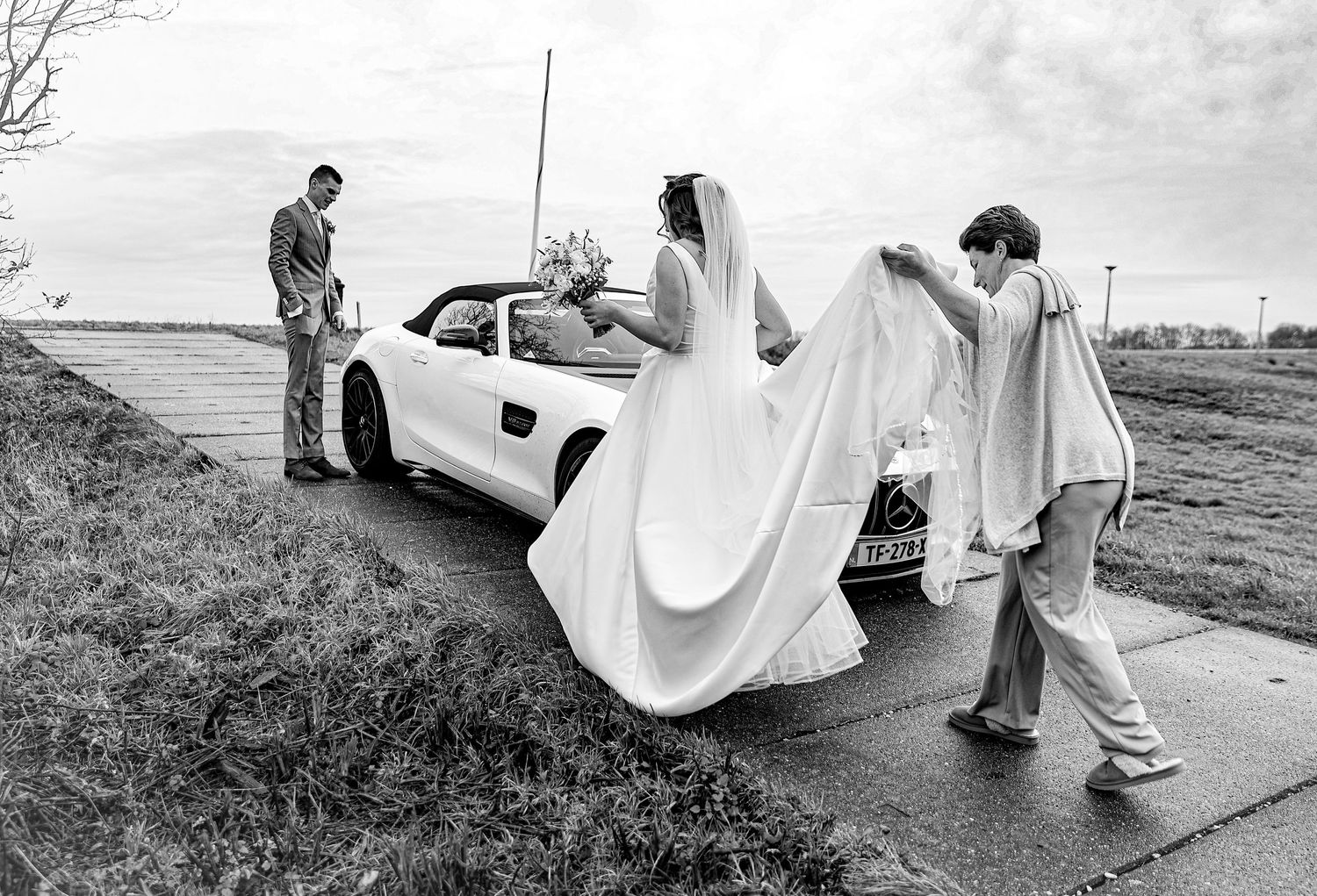 A bride in a flowing white dress being helped into a sports car on a rural road. Wedding photography.