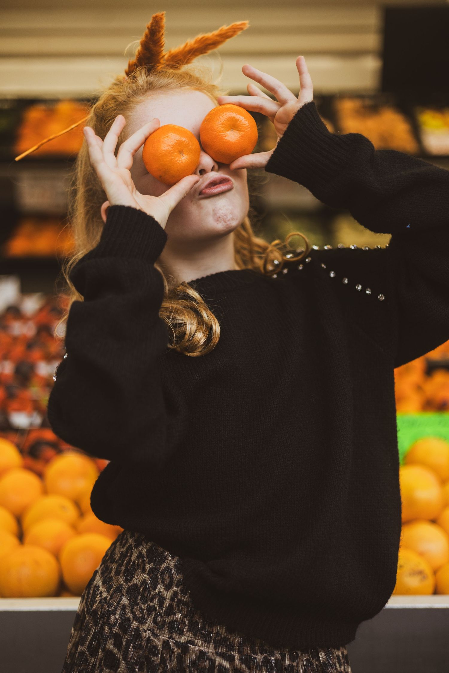 Playful moment at produce stand with oranges held up to eyes while wearing black sweater and patterned skirt.