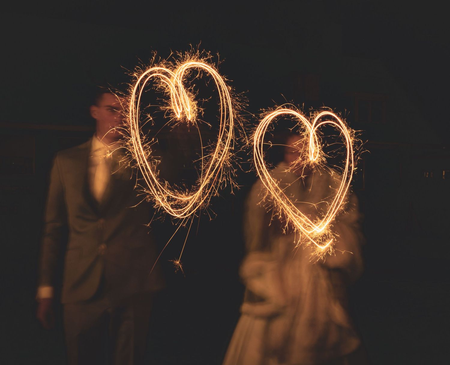 Two glowing sparkler hearts illuminate a dark scene with silhouettes in the background. Movement photography.