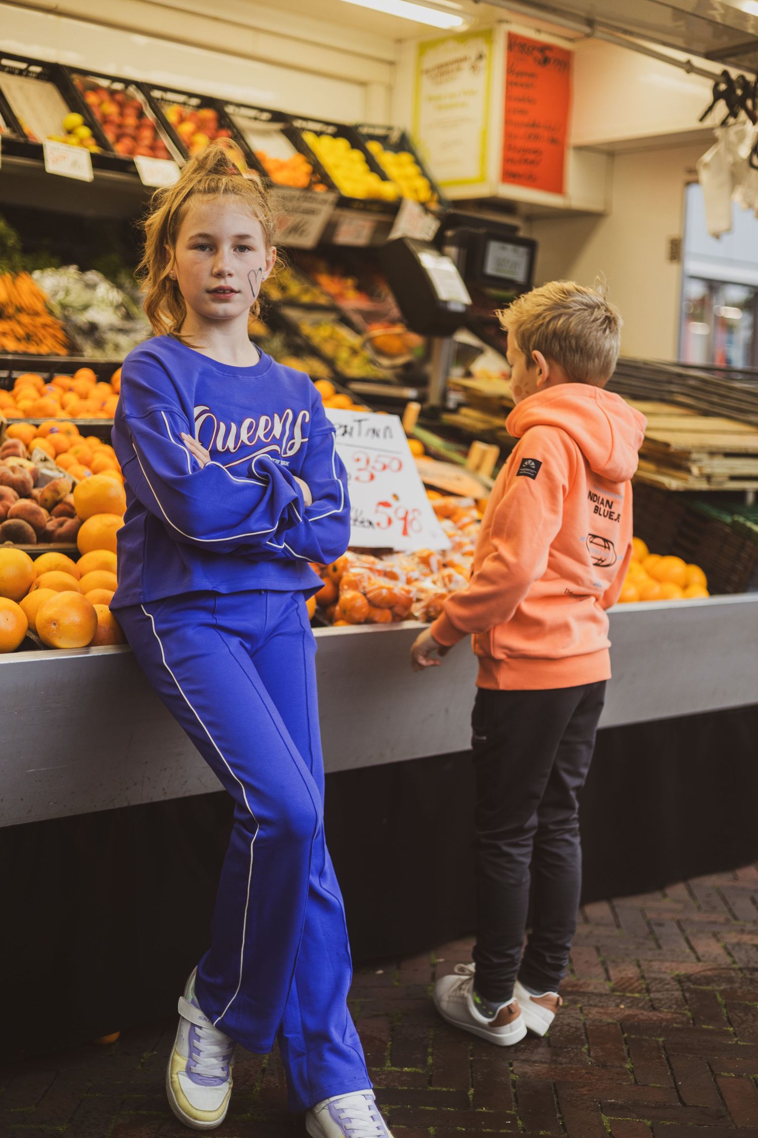Two young people in casual sportswear stand by produce displays at a market with oranges and other fresh fruits.