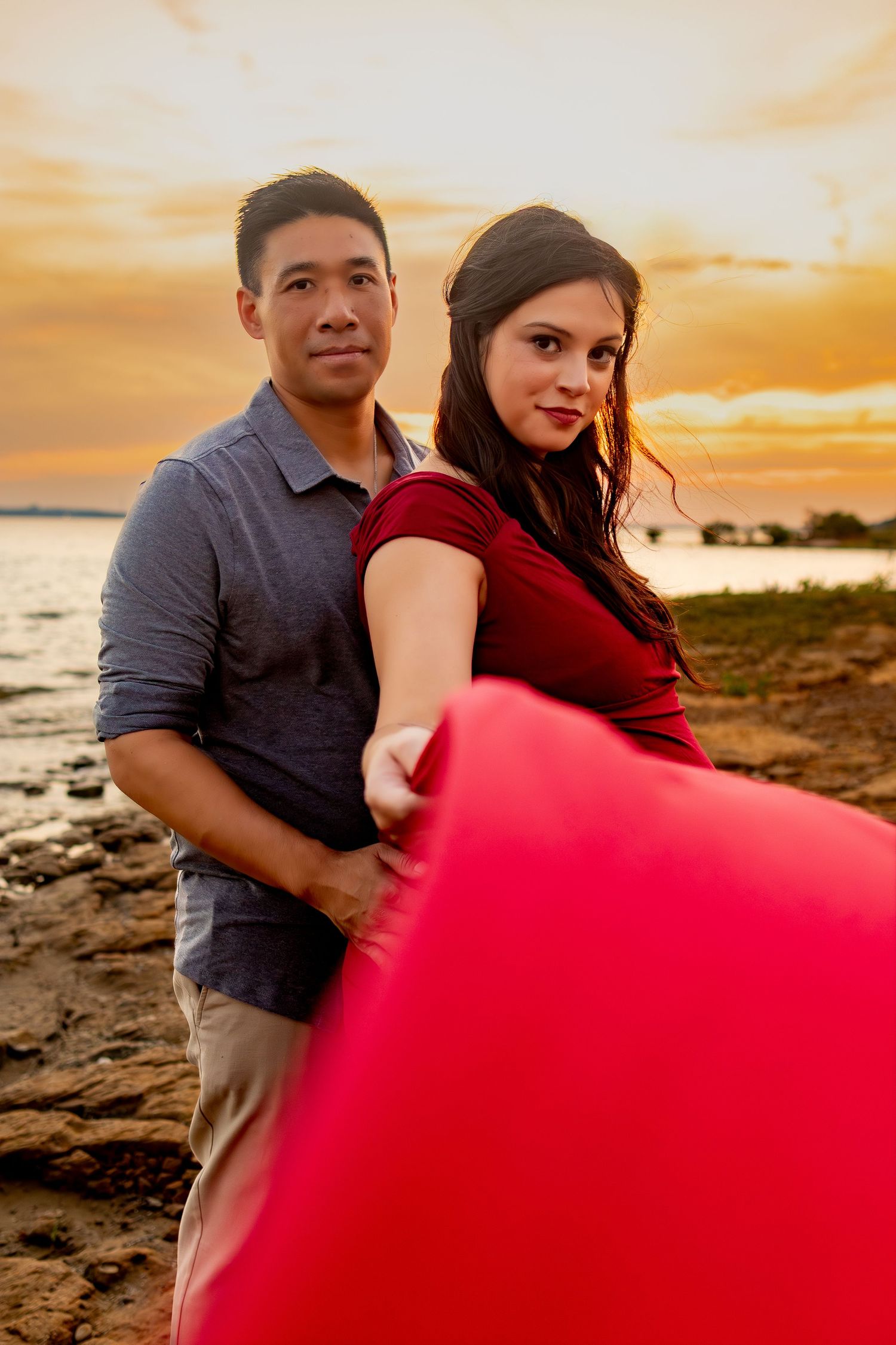 A couple poses with a red blanket on a beach at sunset while waves crash on the rocky shore in the background.