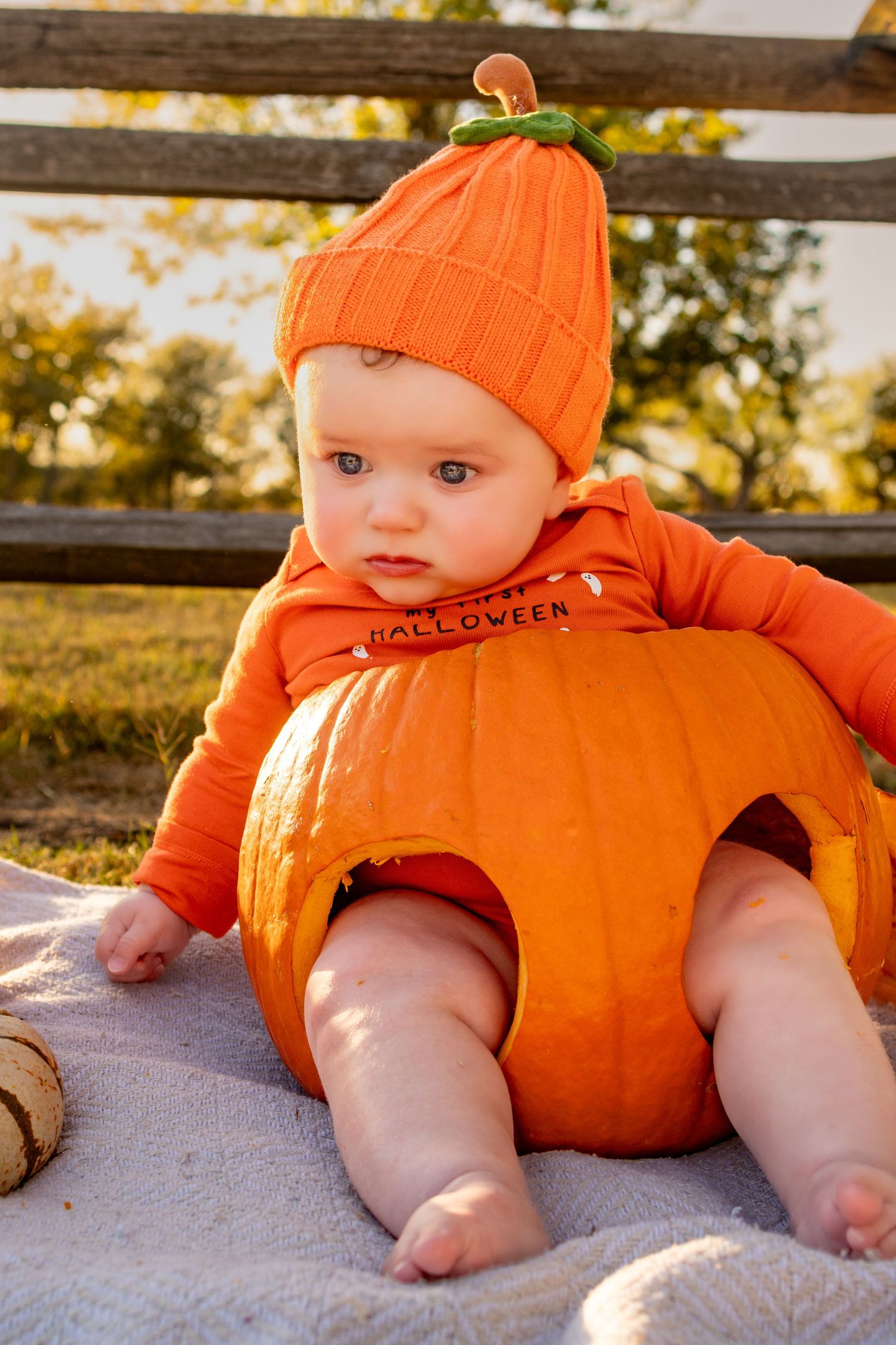 A baby wearing an orange pumpkin costume and hat sits nestled inside a large orange pumpkin outdoors.