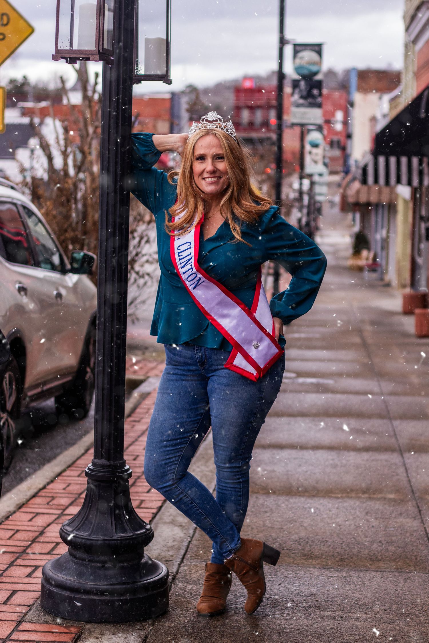 A pageant contestant poses casually against a street lamp wearing a teal blazer and crown on a winter day.