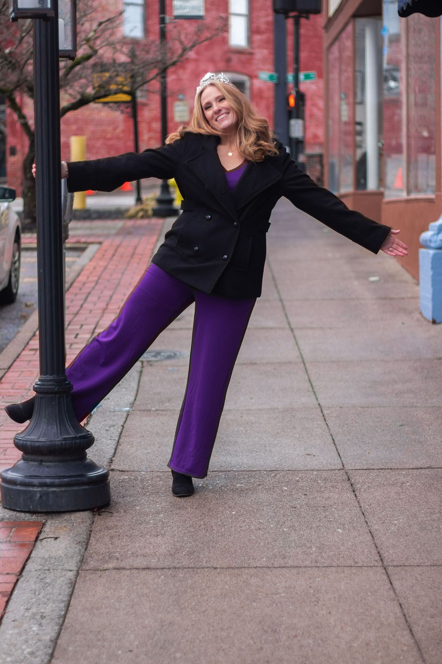 Woman in black coat and purple pants poses playfully with a lamppost on a brick sidewalk.