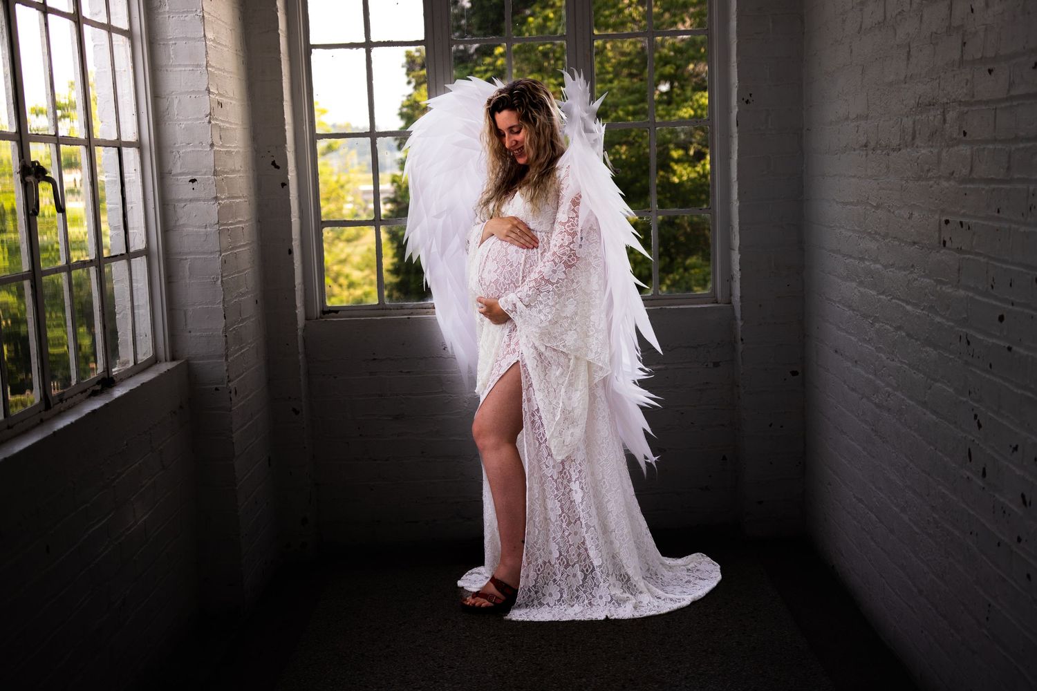 Ethereal maternity photo featuring white lace gown and angel wings in moody industrial setting with natural window light.