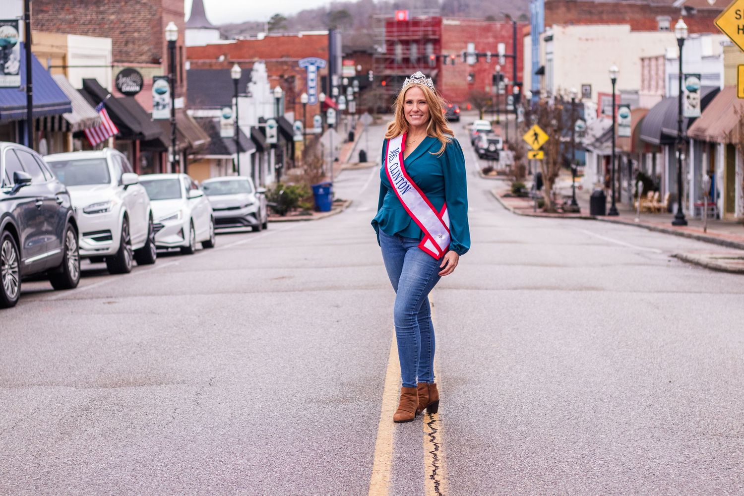 A woman wearing a sash stands confidently in the middle of a small town street wearing blue jeans and teal jacket.