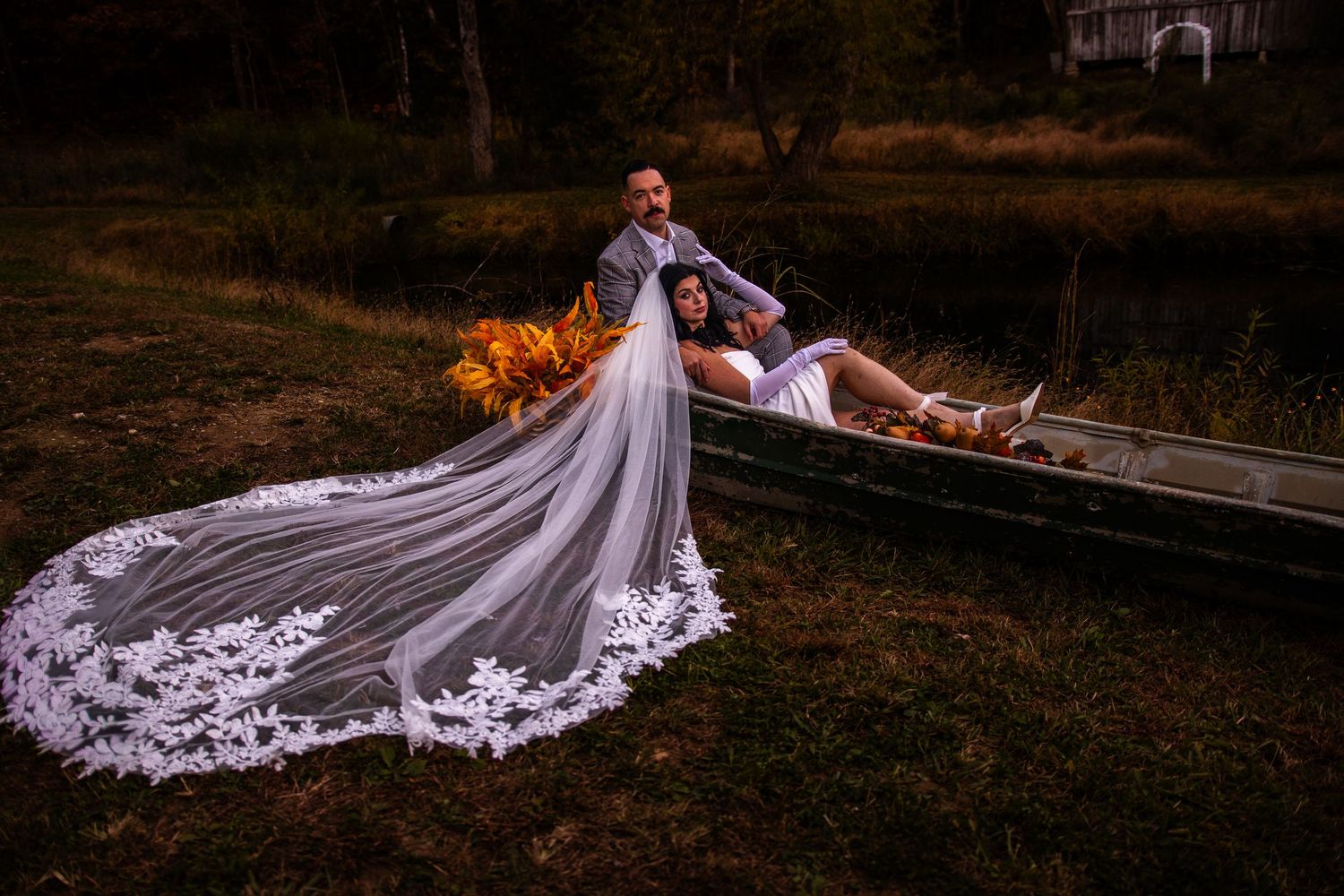 A couple in wedding attire lies together on a rustic path surrounded by autumn foliage and a flowing veil.