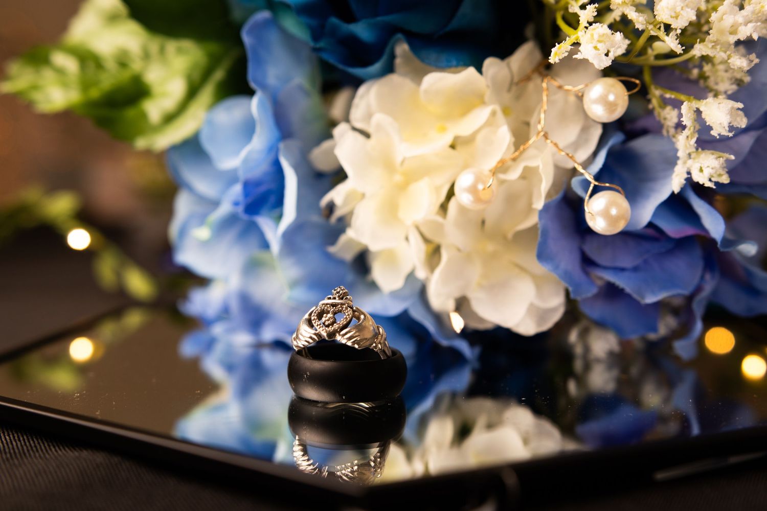 Wedding ring detail shot nestled among blue and white hydrangea blooms with soft bokeh lights in background.
