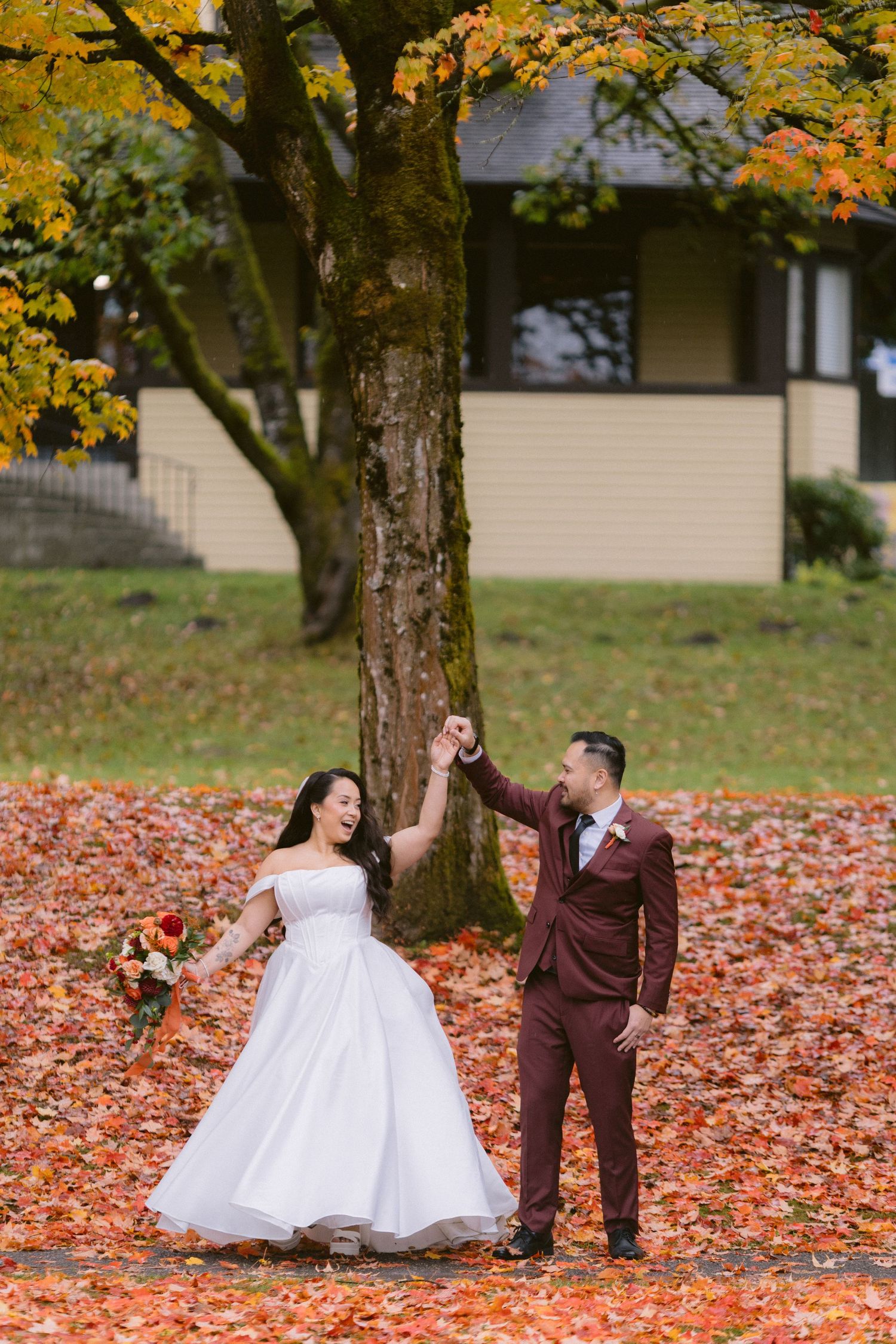 Wedding couple poses playfully among fallen autumn leaves under trees with golden foliage.