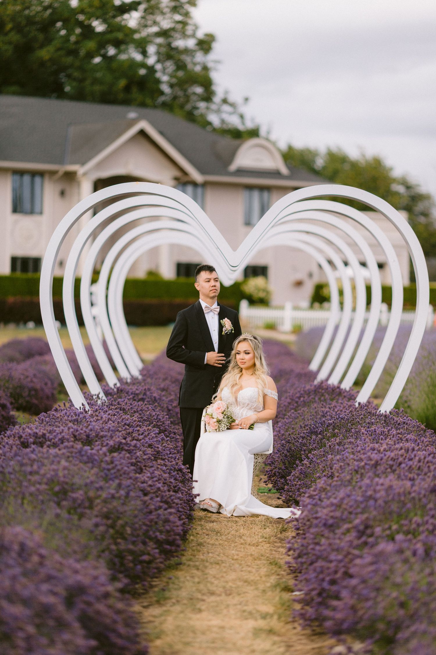 Bride and groom pose in lavender field under white heart arch with house in background.