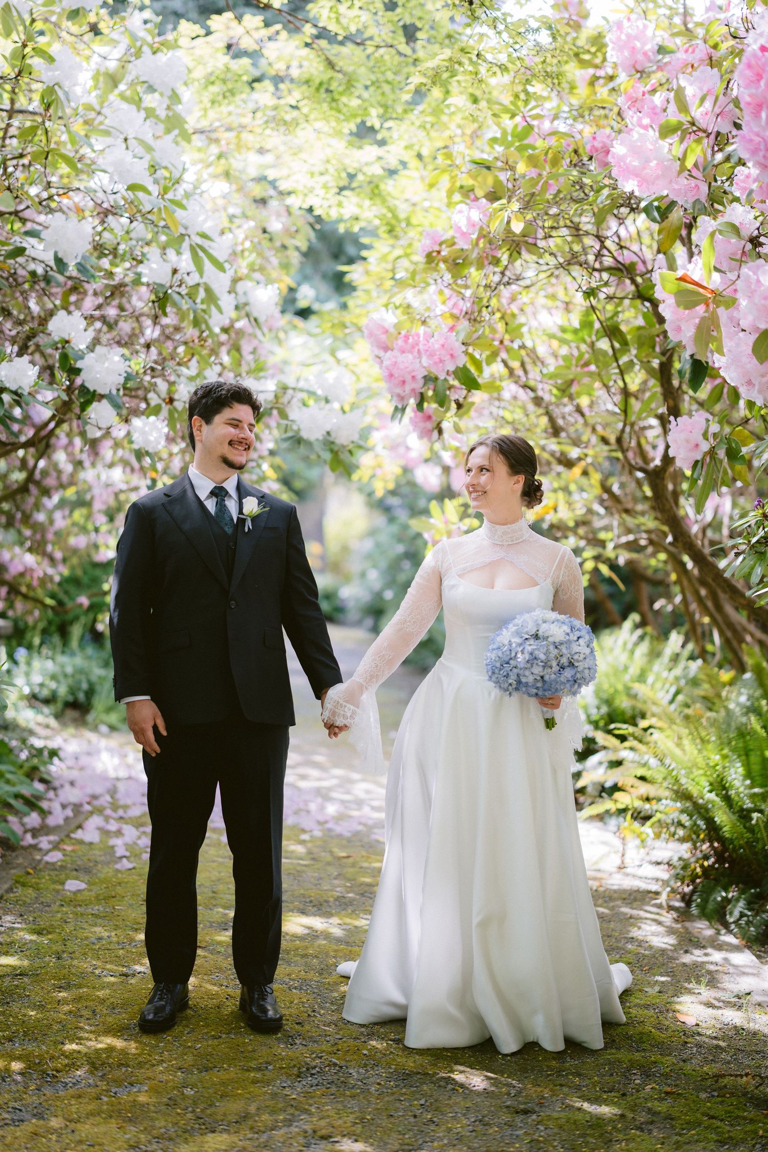 Bride and groom hold hands walking through a blooming garden path with pink and white flowers on their wedding day.