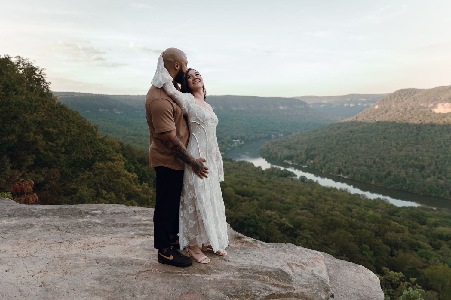 A couple embraces on a scenic mountain overlook with a winding river valley stretching into the distance.