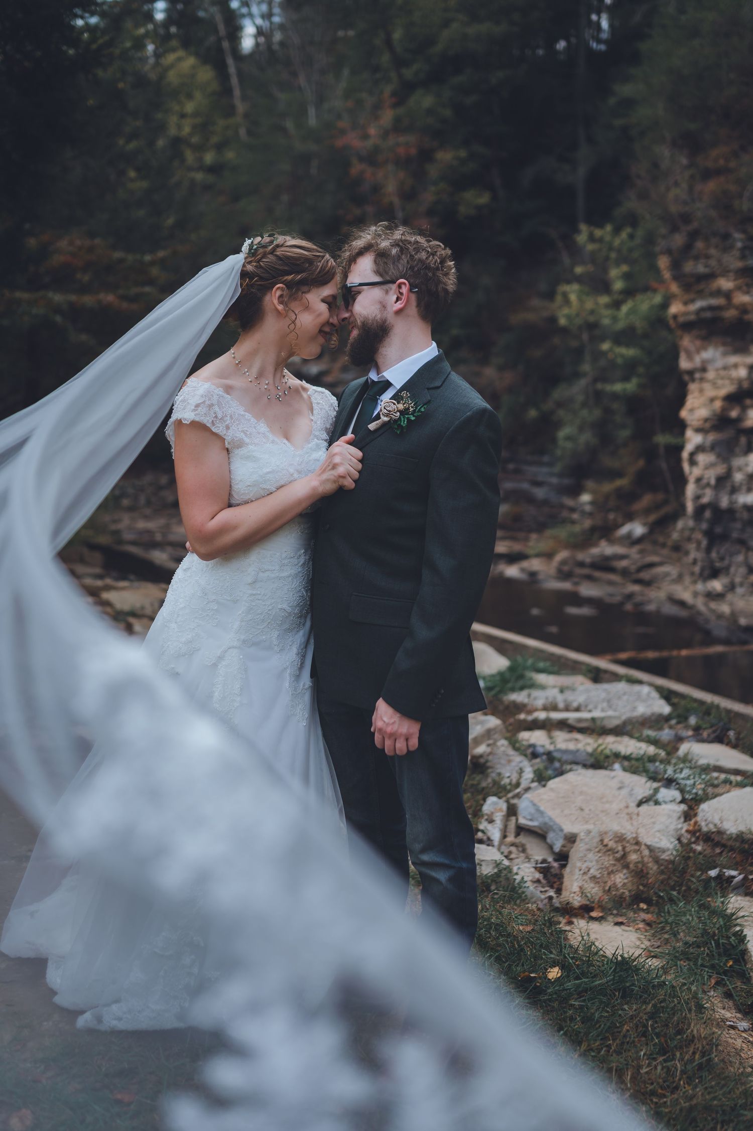 Bride and groom share romantic moment outdoors, veil flowing in wind, rocky creek backdrop, autumn wedding portrait.