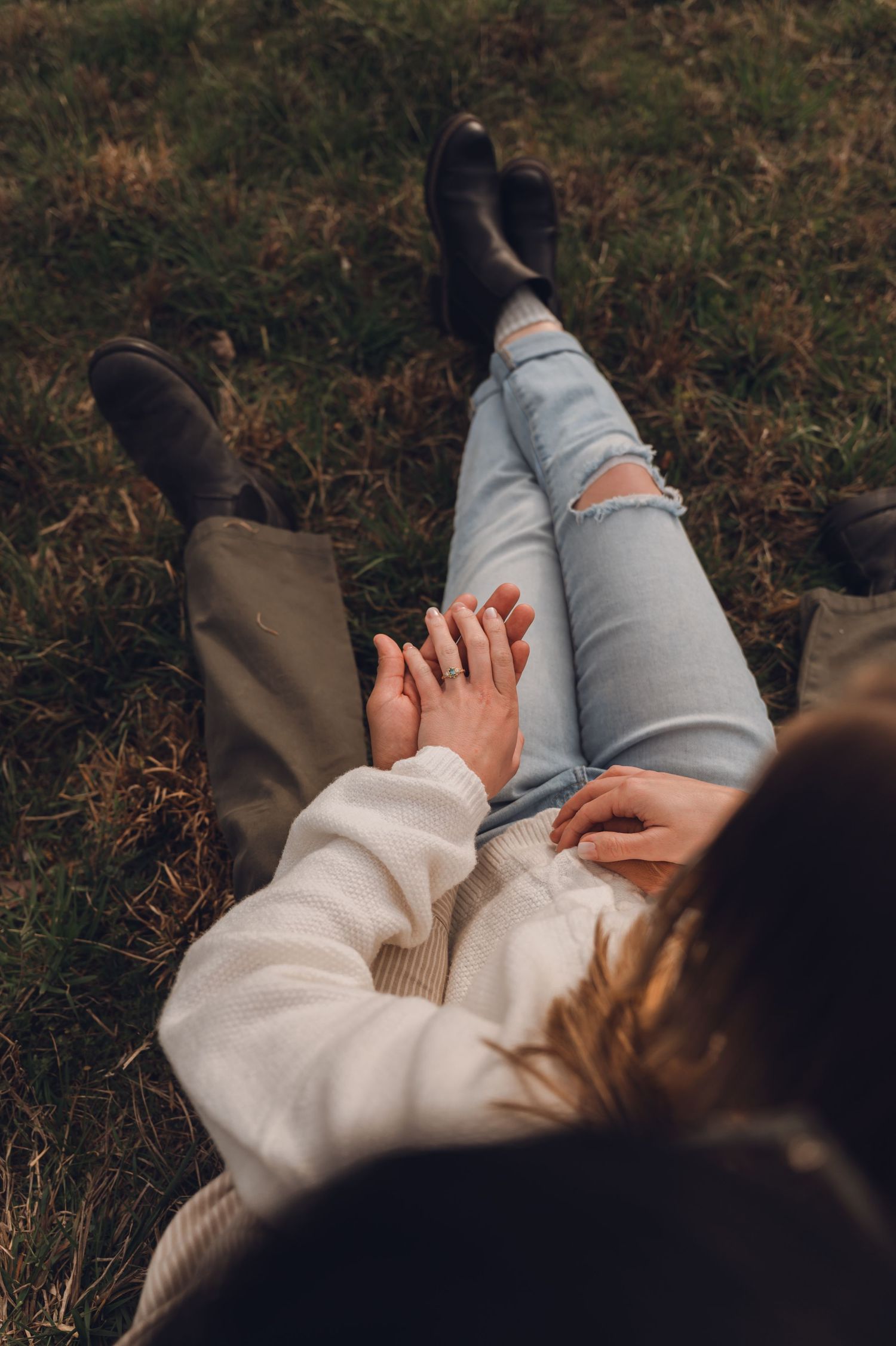 Couple holding hands while lying on grass, wearing jeans and boots in an autumn outdoor setting.