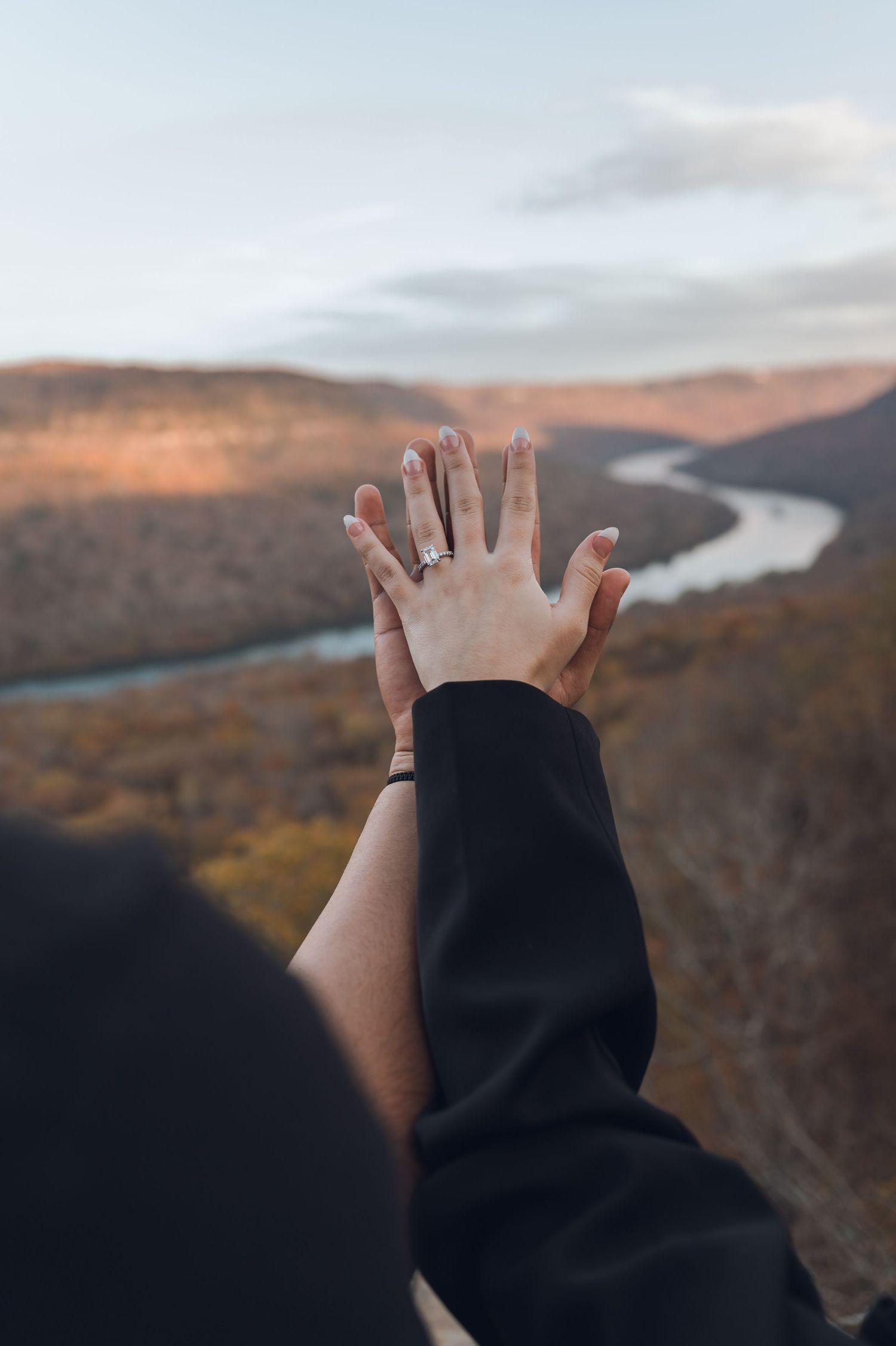 A hand reaches out towards a winding river valley during sunset, with mountains and autumn foliage in the background.