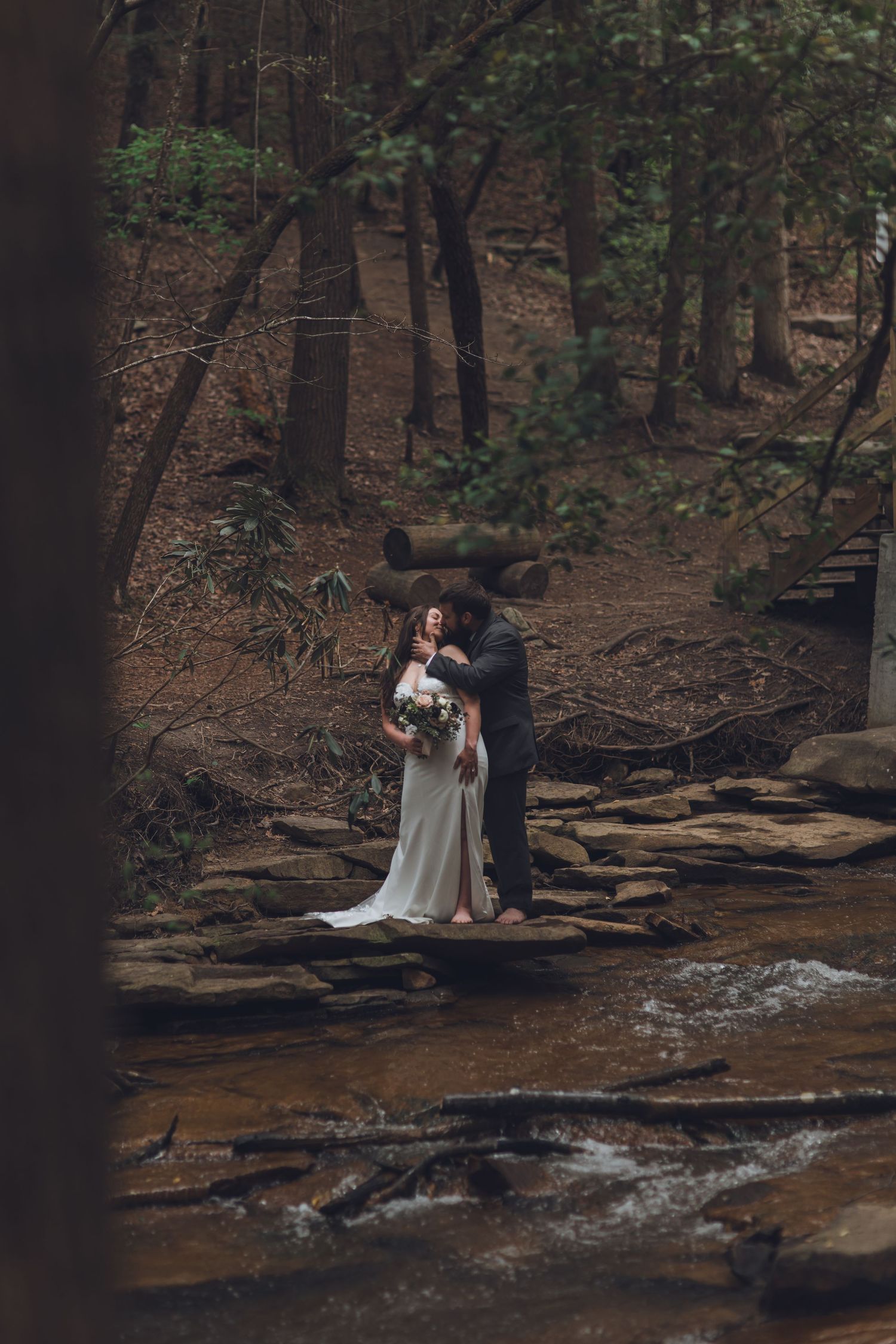 Bride and groom embracing on rocky creek bed surrounded by lush forest trees during outdoor woodland wedding ceremony.