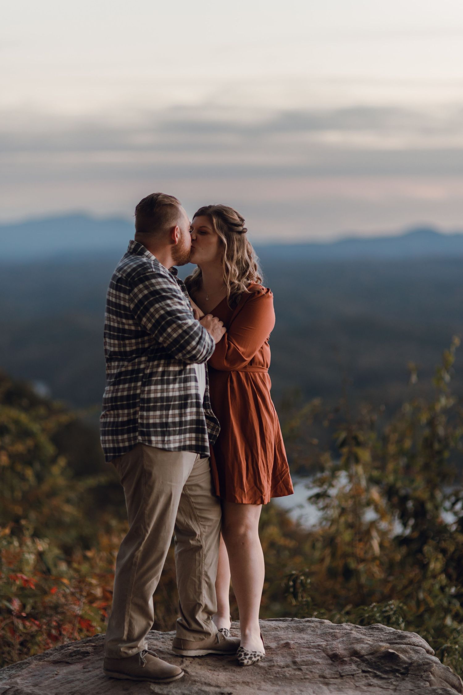 Couple kissing on a rocky overlook with mountain views at sunset, man in plaid shirt, woman in rust orange dress.