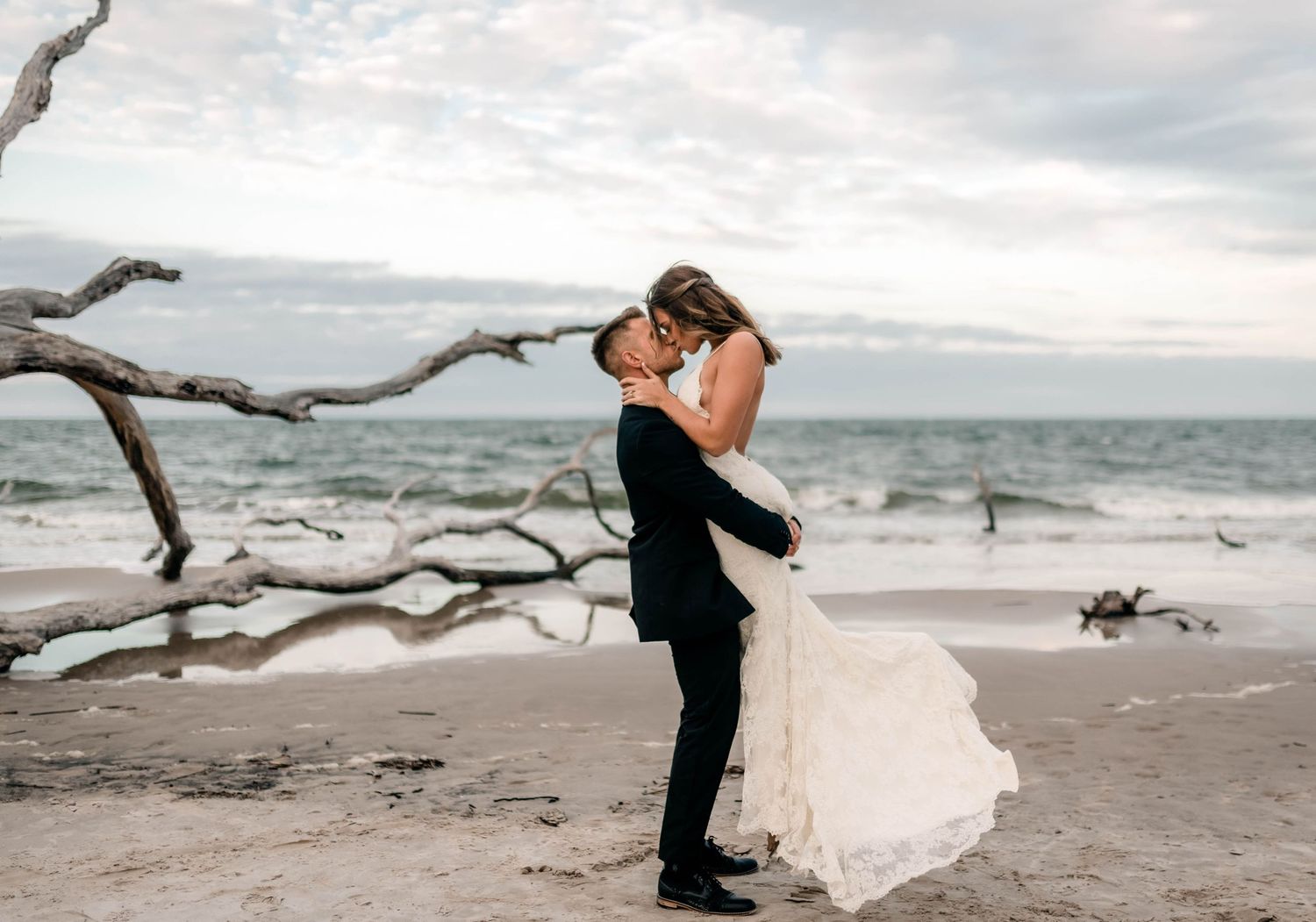 A romantic couple embraces on a beach with driftwood and ocean waves in the background at sunset.