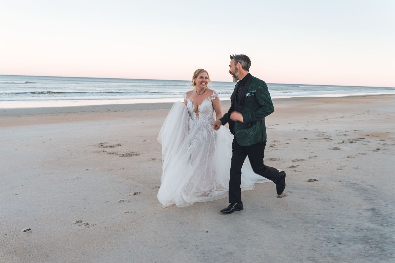 A couple in wedding attire walks together along a serene beach at sunset, creating romantic silhouettes in the sand.