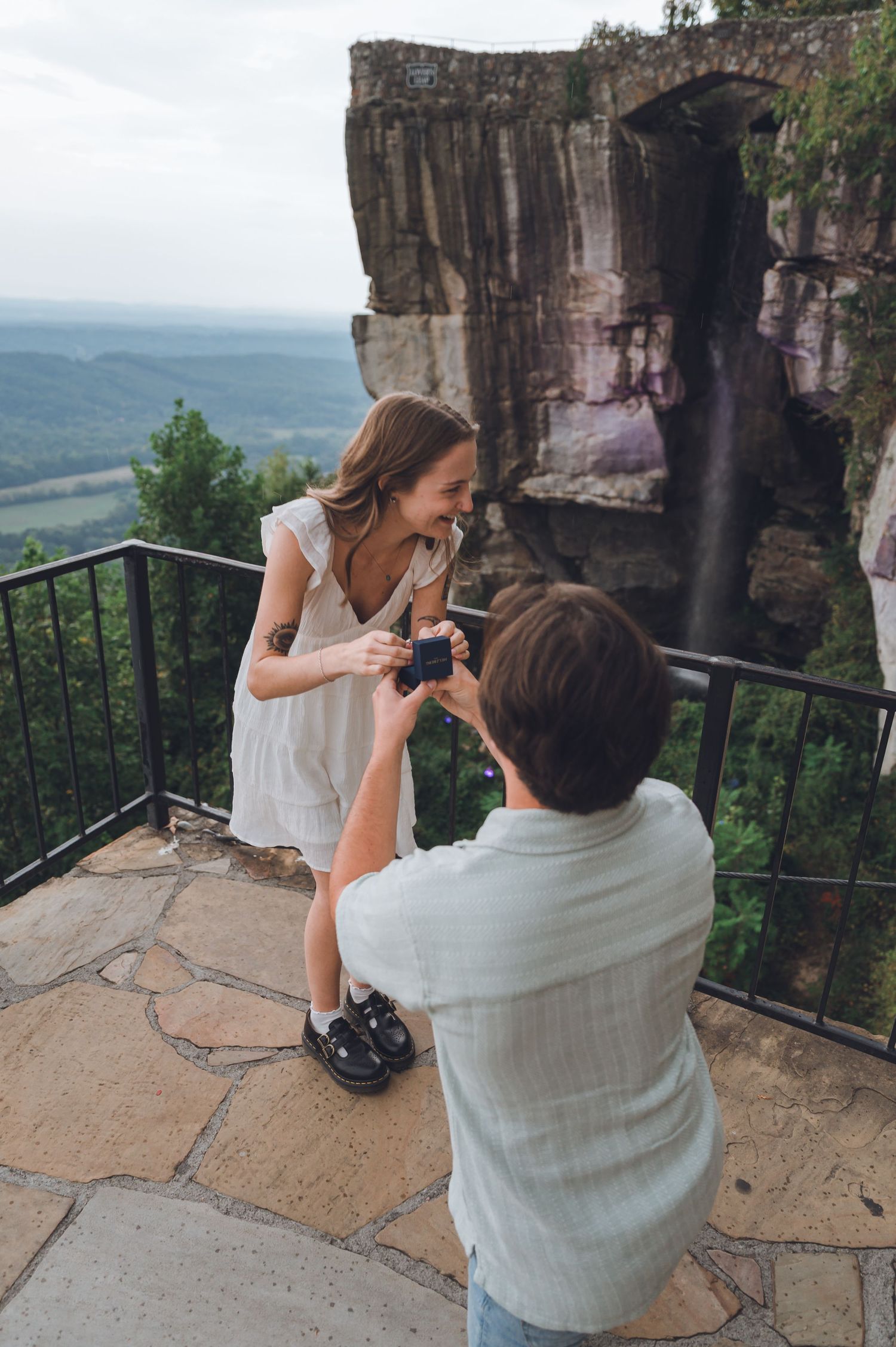Man proposing to woman on scenic overlook with waterfall and valley views in background.