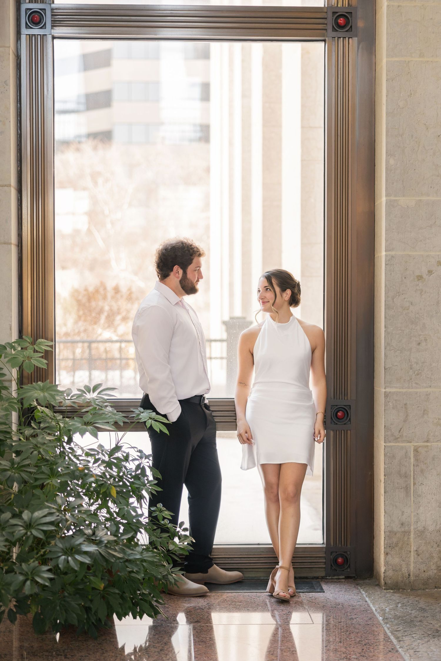 Couple in formal attire poses for romantic engagement photos in elegant building hallway with natural lighting.