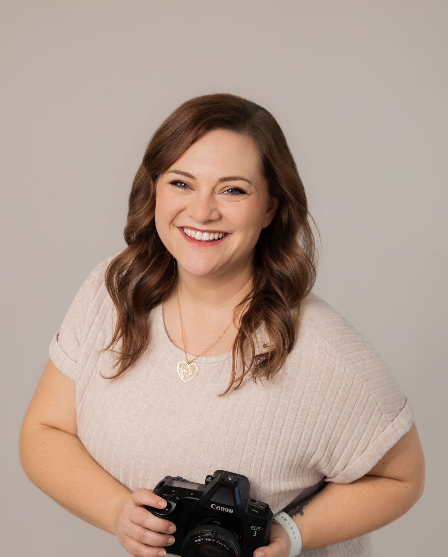 Professional photographer in light beige sweater smiles warmly while holding a DSLR camera.