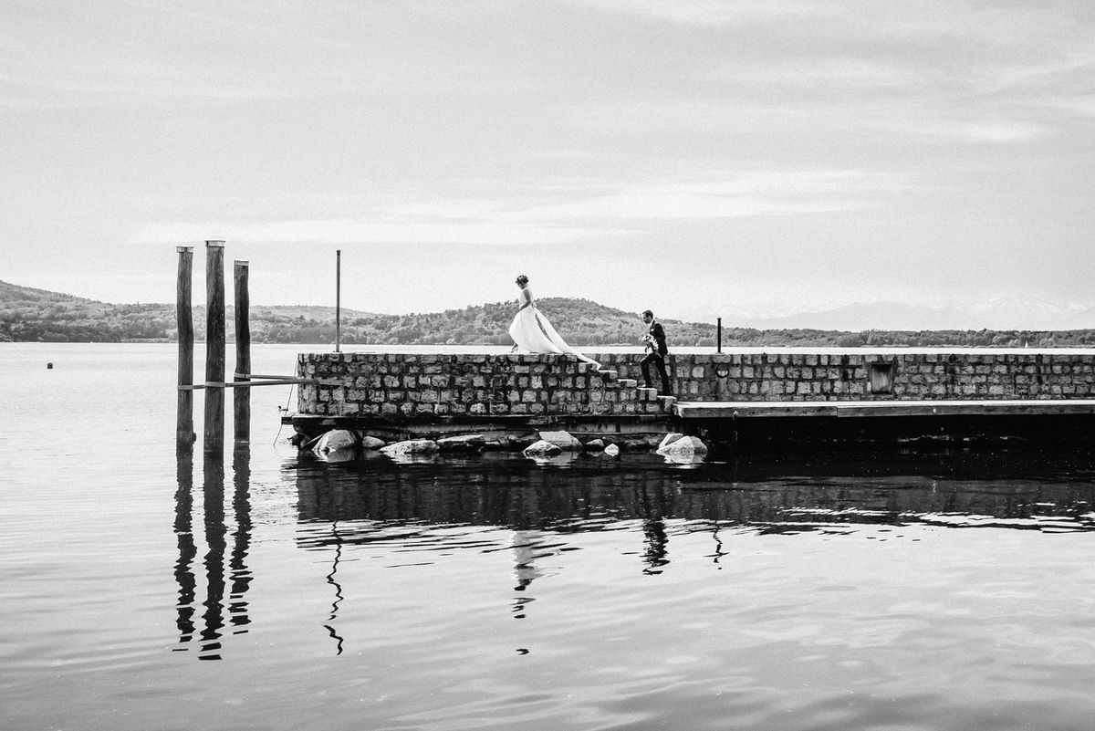 Due sposi su un molo di un lago con pali di legno e riflessi d'acqua calma in fotografia in bianco e nero.