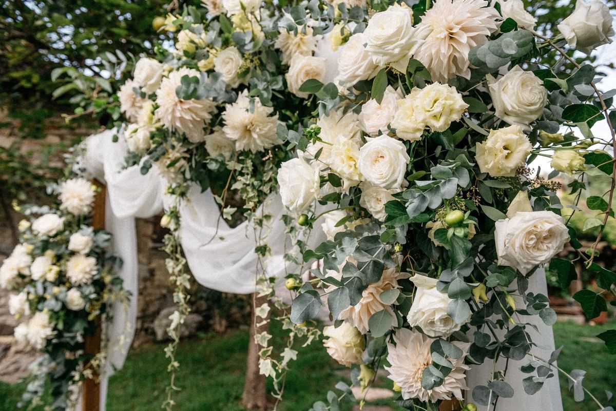 Eleganti rose bianche e vegetazione creano una romantica installazione ad arco floreale per una cerimonia di matrimonio in giardino.