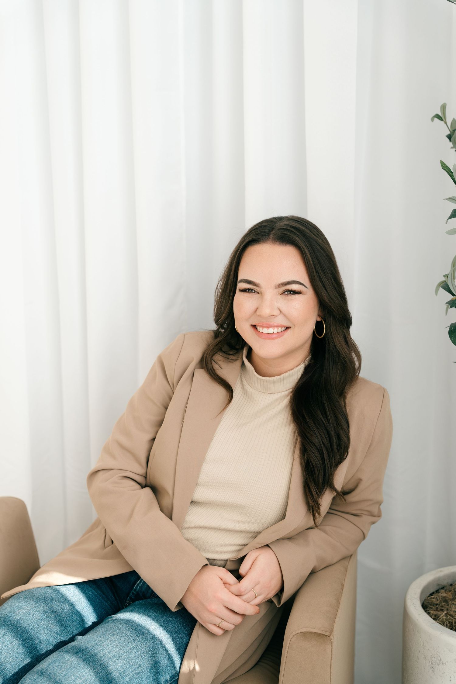 Woman in tan blazer and turtleneck sitting by white curtains.