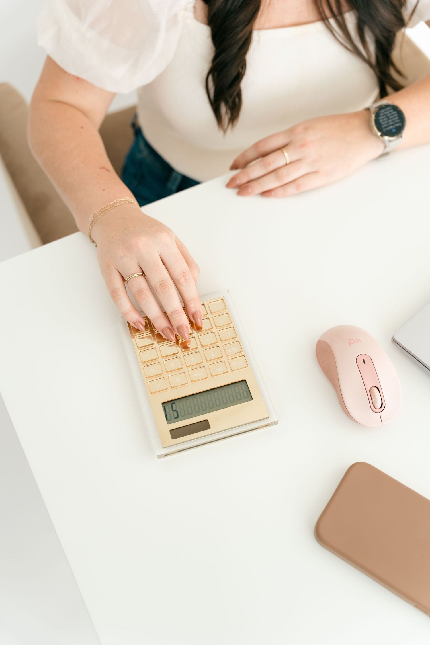 Person using gold calculator and pink mouse on white desk with phone.