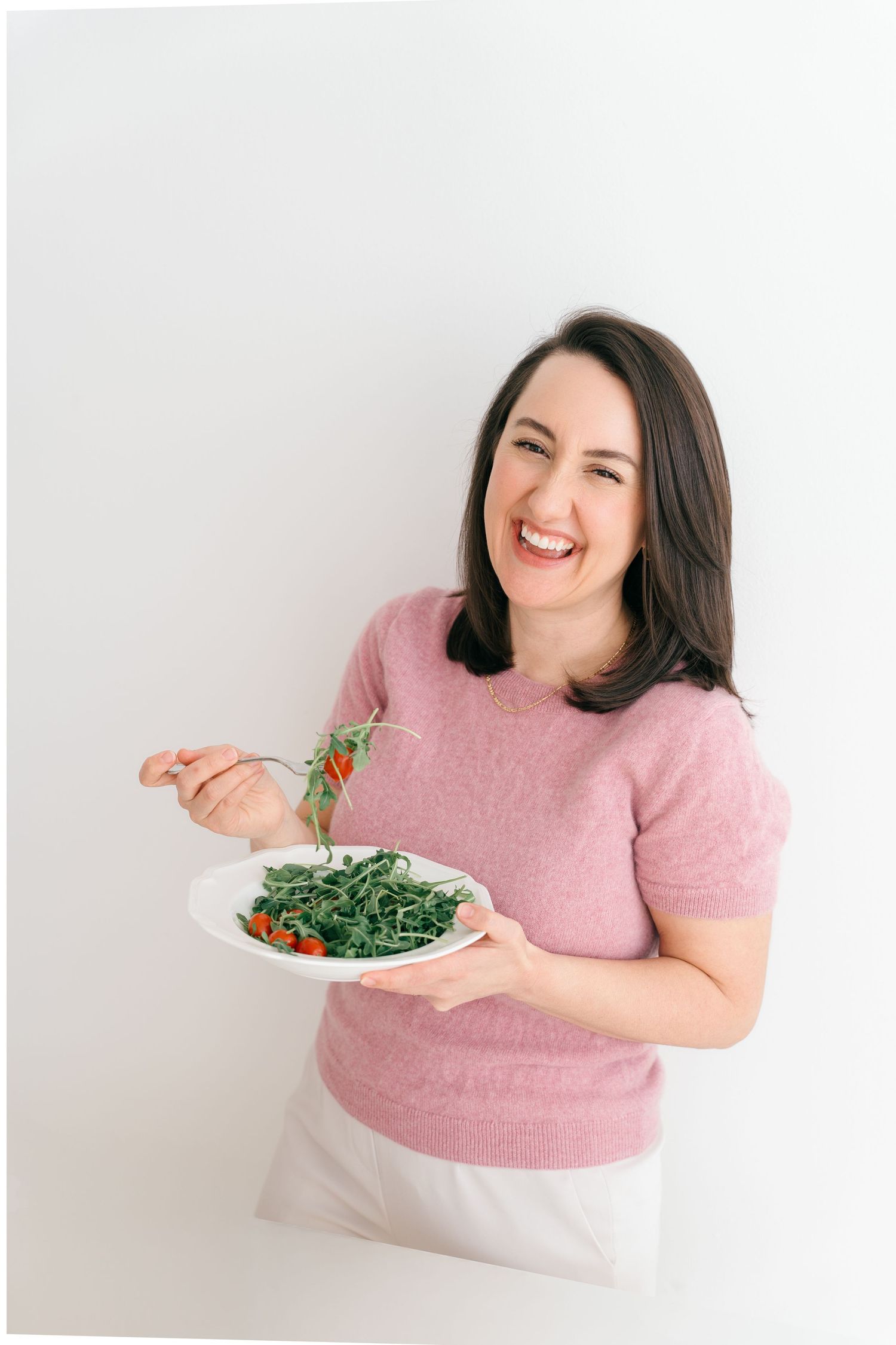 Woman in pink sweater laughing while holding plate of fresh green salad against white wall background.