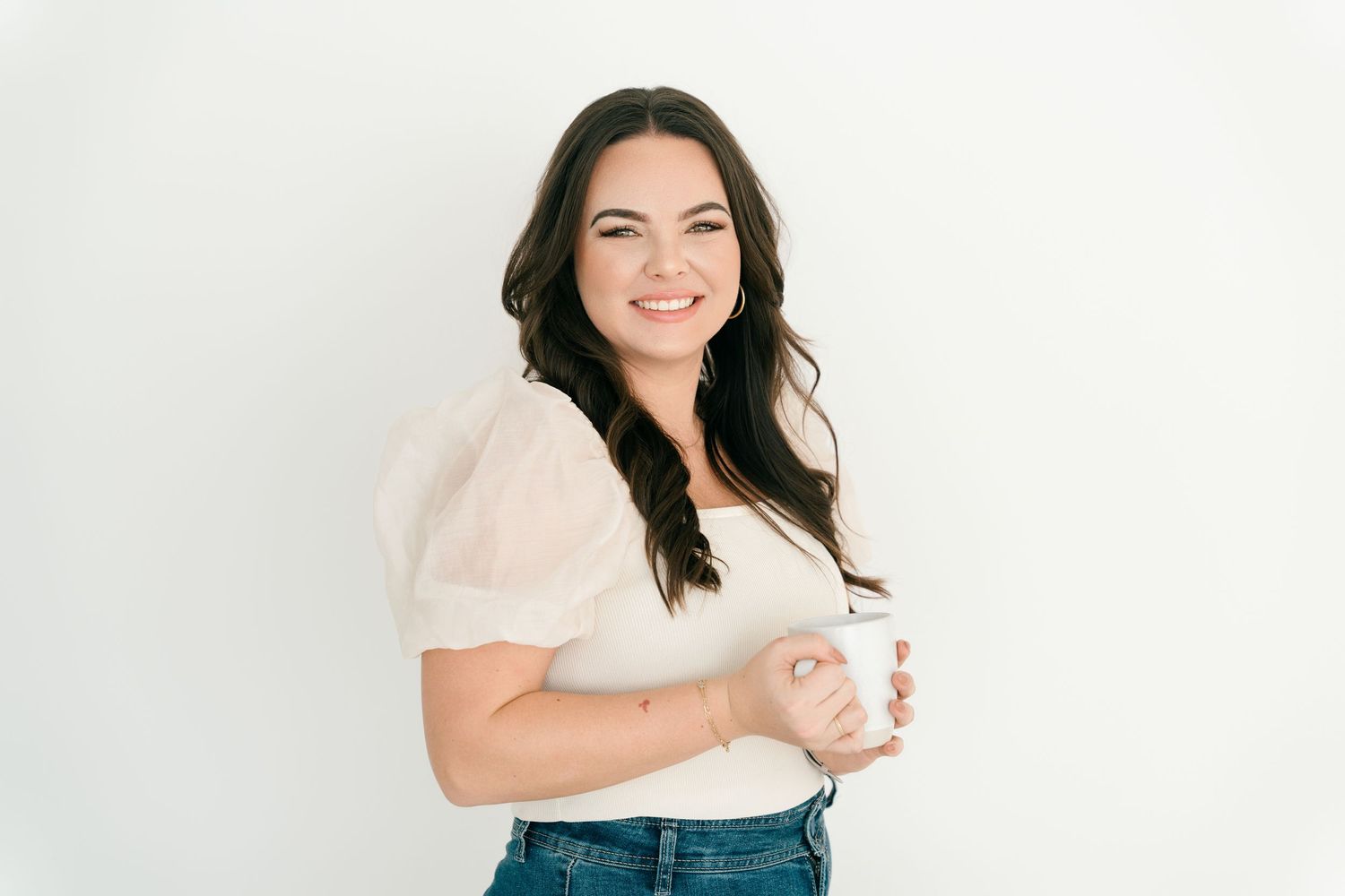 Woman in white blouse holding coffee mug against white wall.