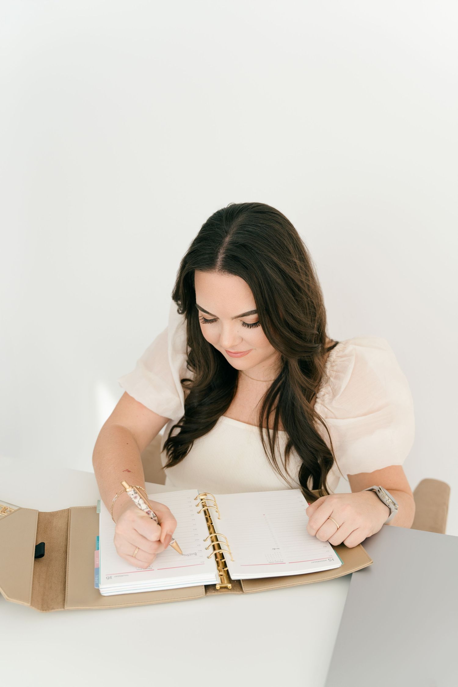 Woman writing in planner at white desk with calculator and office supplies.