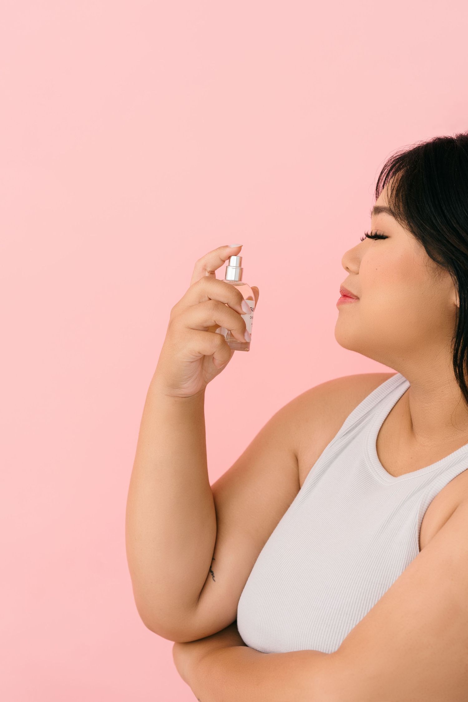 Person in white tank top spraying perfume against pink background in a profile view.