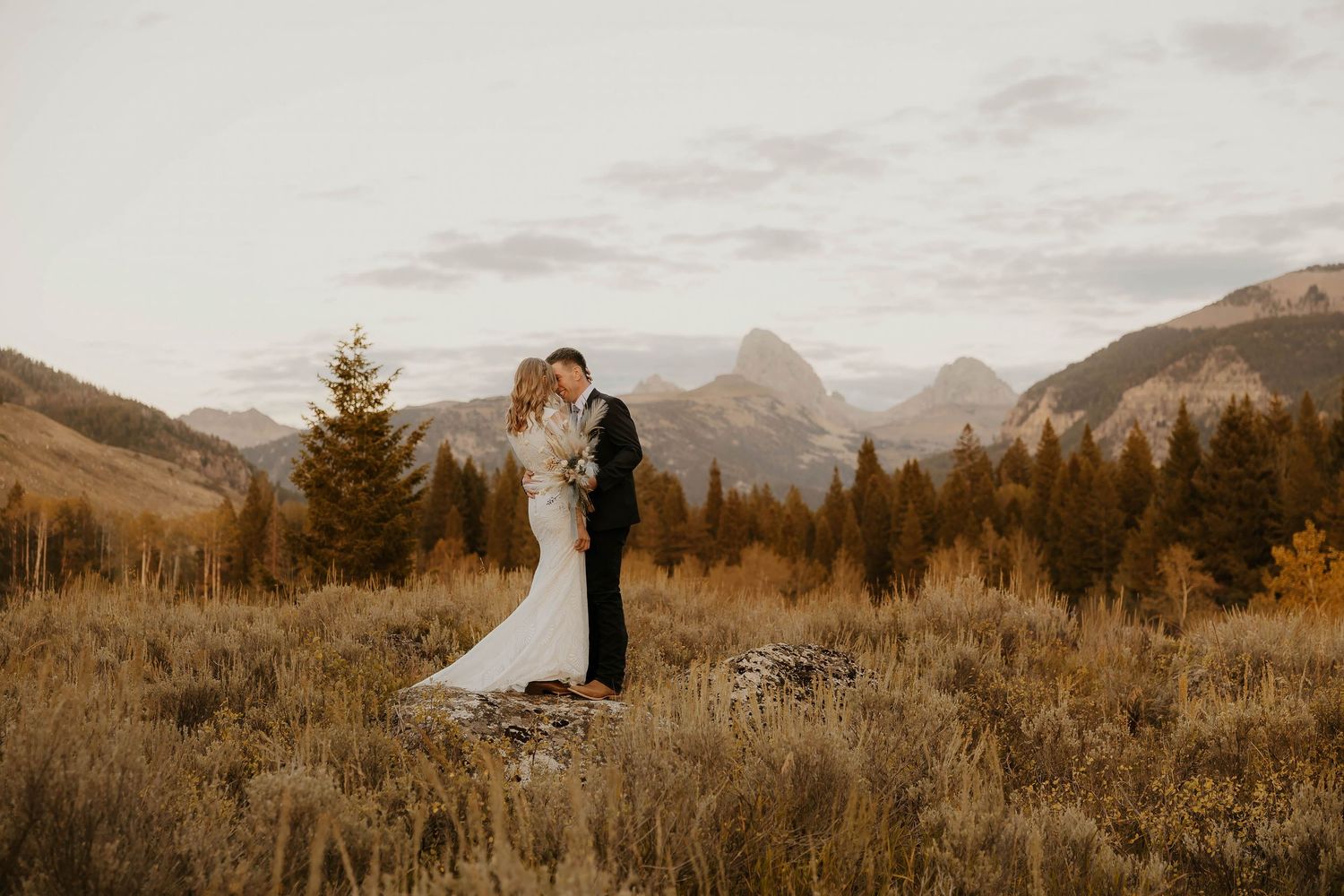 Bride and groom embrace in golden meadow with mountain backdrop during outdoor fall wedding ceremony.