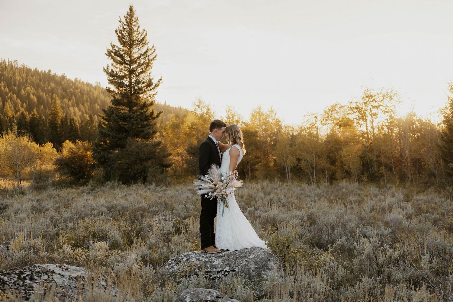 Bride and groom kissing in a golden meadow surrounded by autumn trees at sunset, bride holding a boho bouquet.