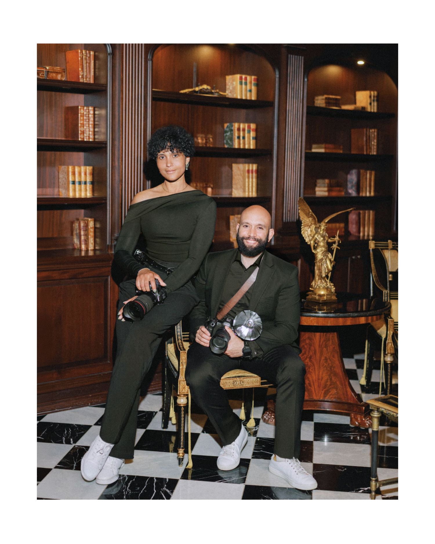 Two photographers in matching olive outfits pose with cameras in a sophisticated library setting.