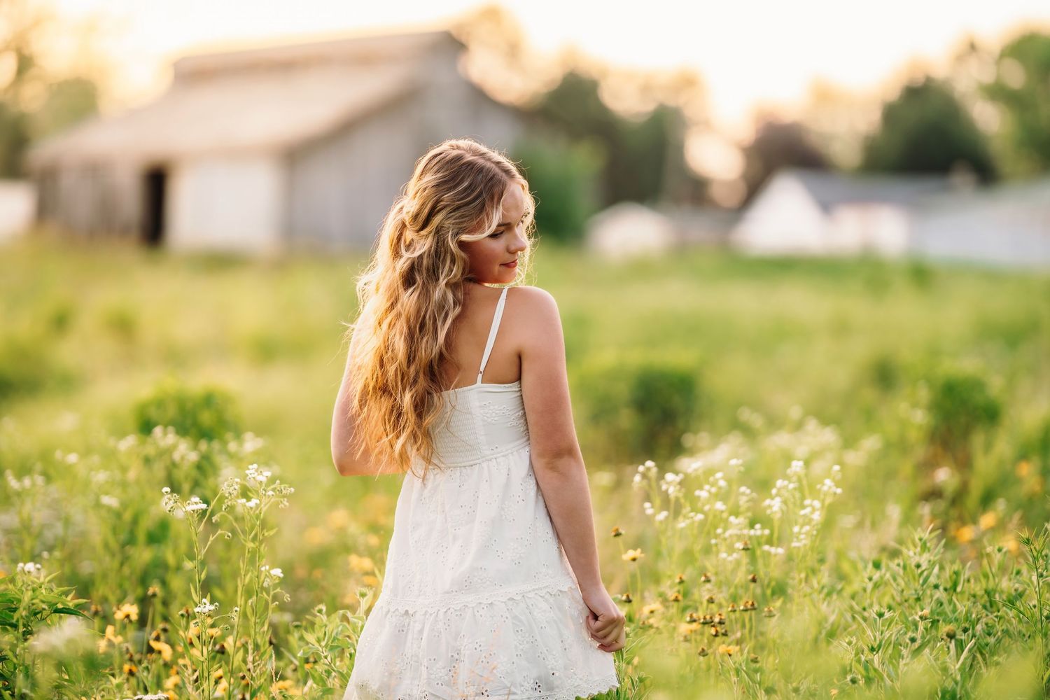 A person in a flowing white dress walks through a sunlit meadow of wildflowers at golden hour. Senior photography Cincinnati.